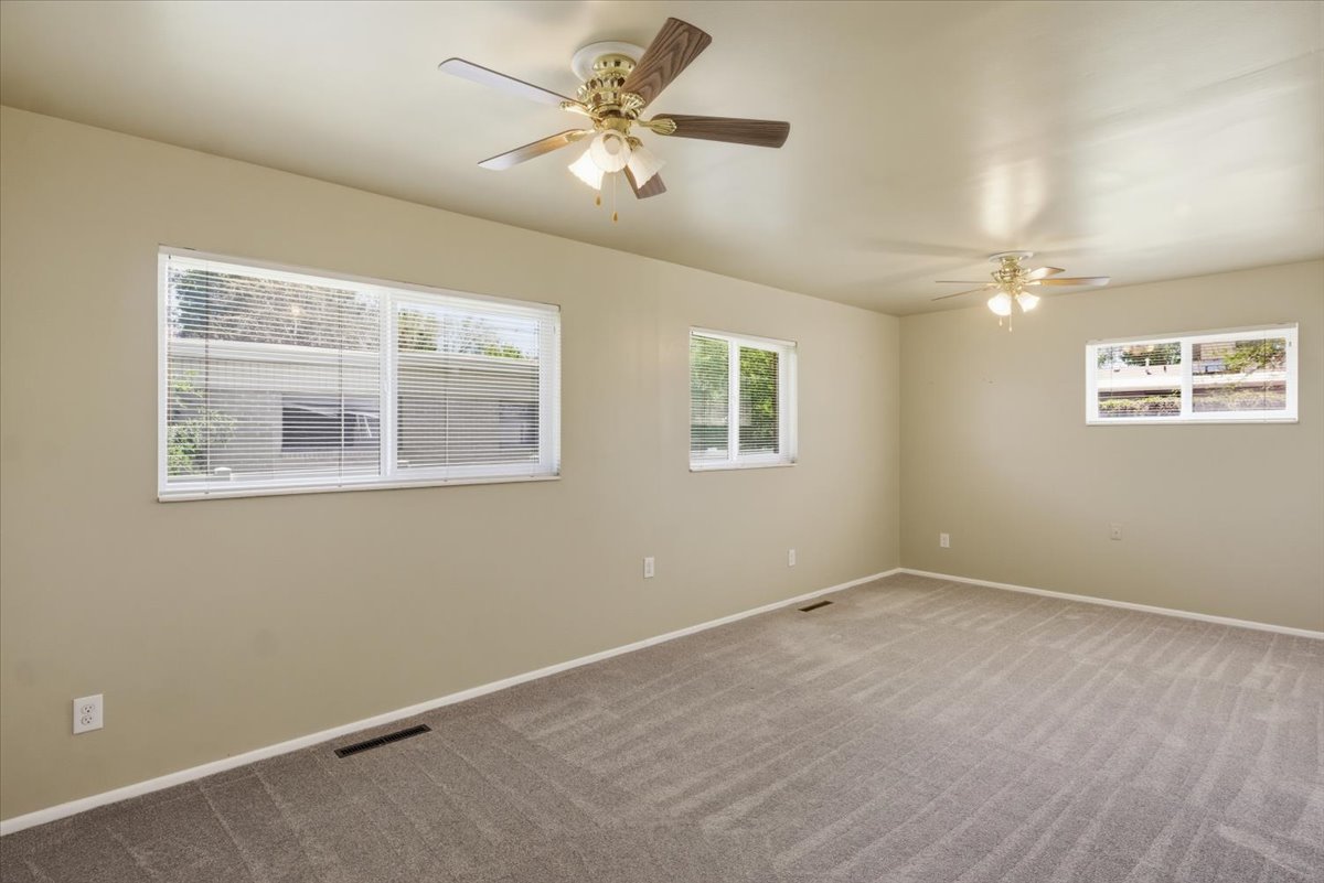 Spacious room featuring neutral-toned walls, ceiling fans with integrated lighting, multiple windows with blinds, and patterned carpet flooring