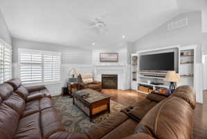 Living area featuring wood-finish flooring, a vaulted ceiling, a gas fireplace with a white mantel, custom built-in shelving, and plantation shutters
