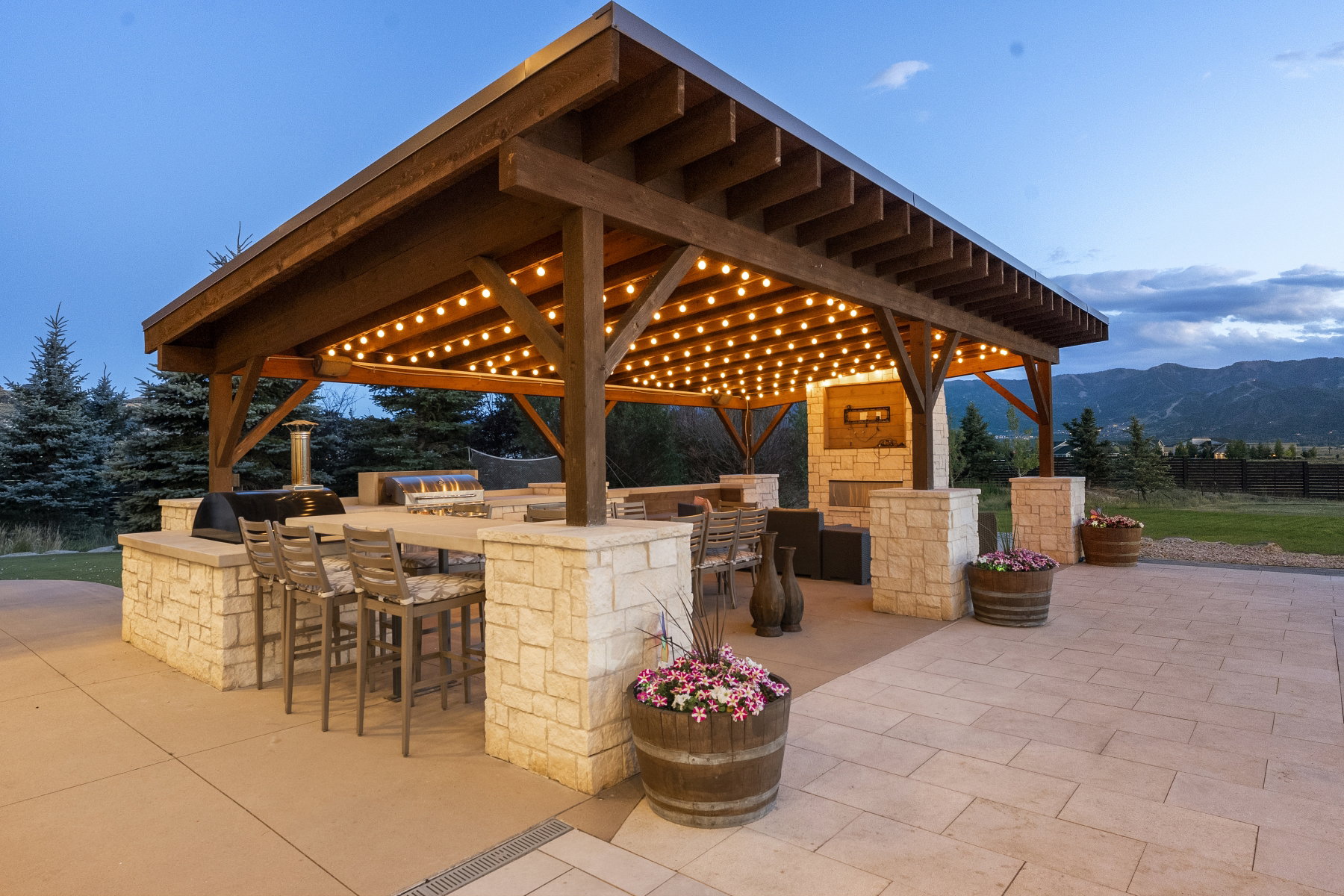 Expansive outdoor pavilion featuring a wood-beam ceiling with string lighting, a stone-clad outdoor kitchen, a built-in fireplace, and a large stone countertop bar