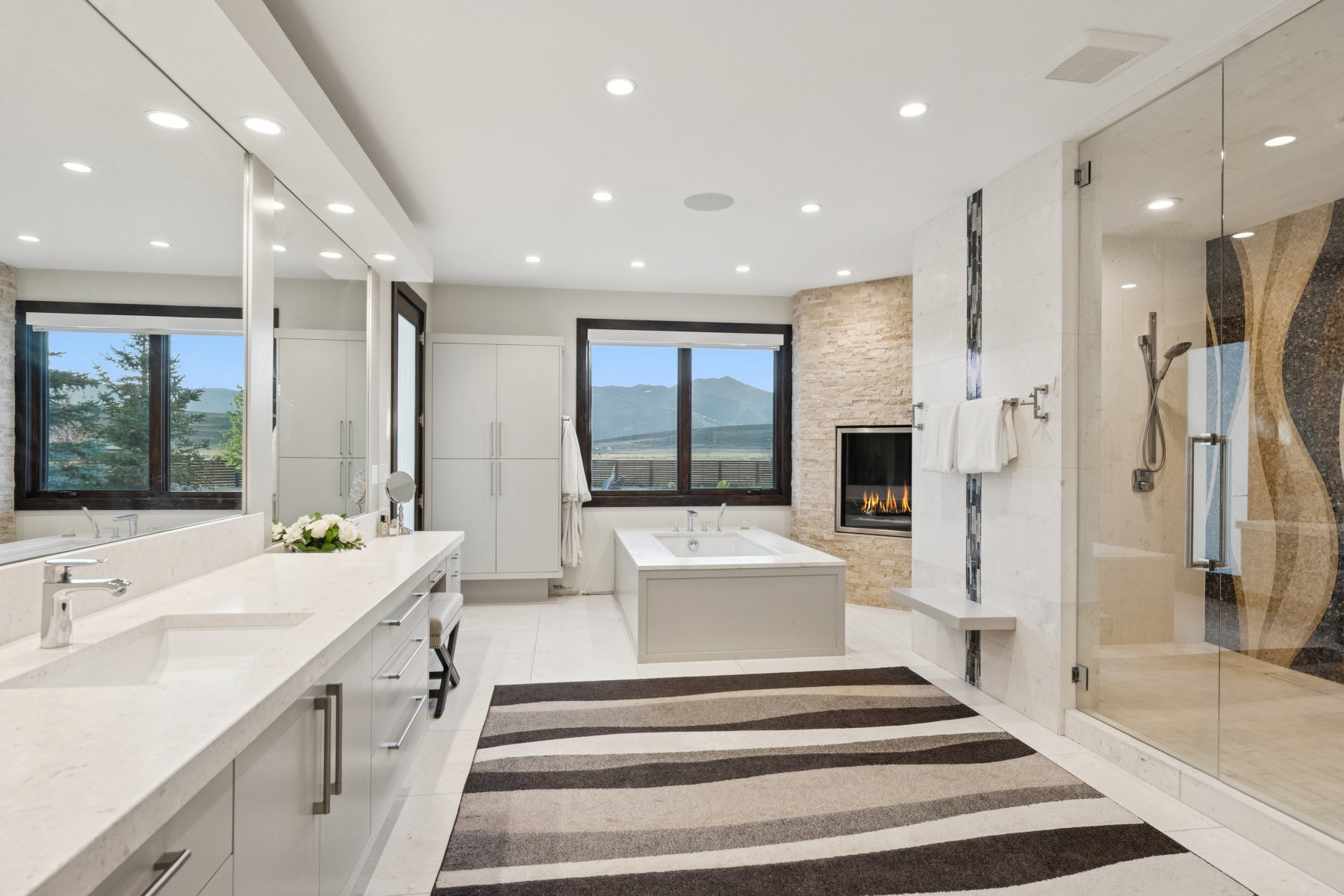 Spacious bathroom featuring a dual vanity with undermount sinks and extensive countertop space, an integrated fireplace with stone surround, a soaking tub, and a glass-enclosed shower with a decorative tile wall