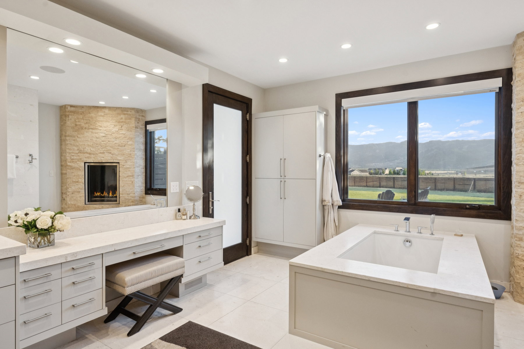 Spacious bathroom featuring a built-in soaking tub, stone-clad fireplace, integrated vanity with ample storage, large mirror, and panoramic mountain scenery