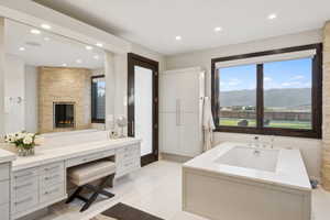 Spacious bathroom featuring a built-in soaking tub, stone-clad fireplace, integrated vanity with ample storage, large mirror, and panoramic mountain scenery