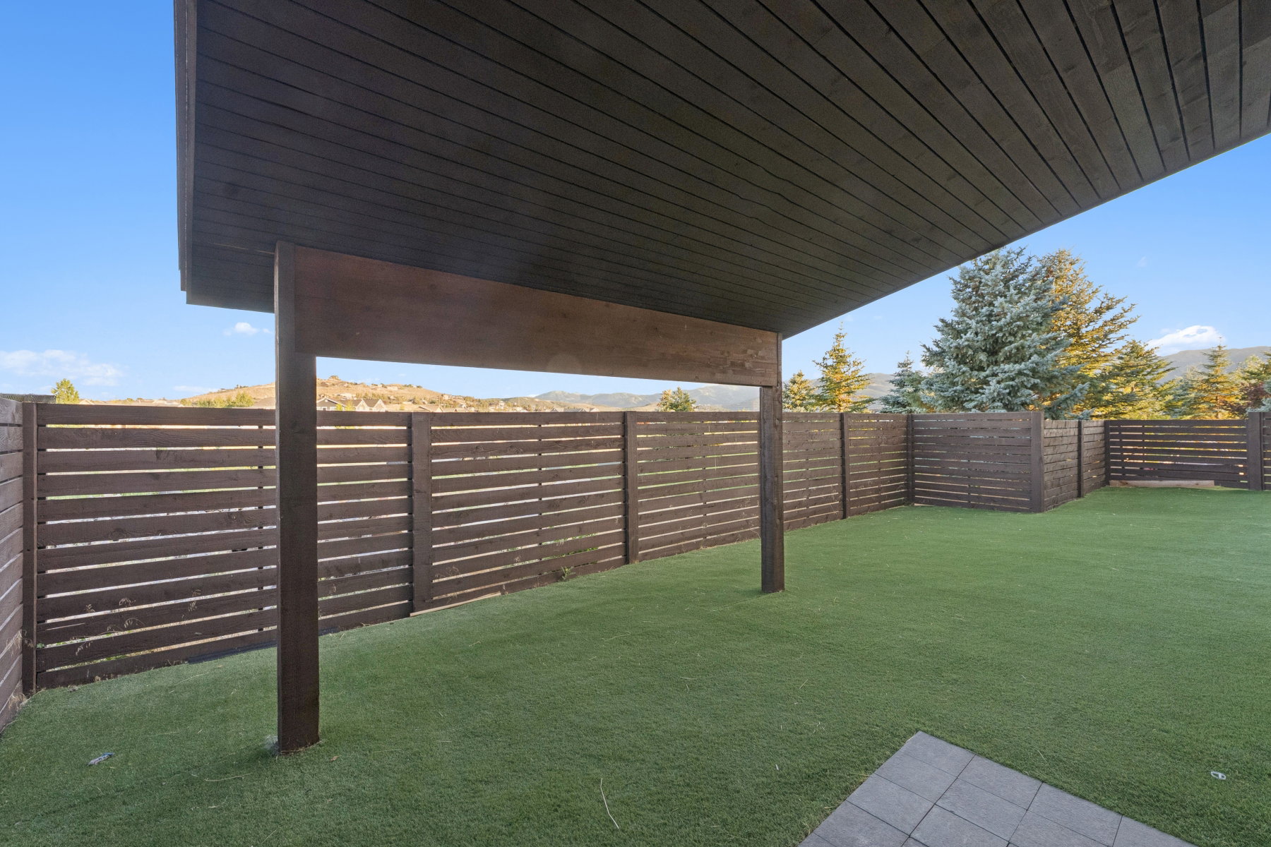 Expansive outdoor area featuring a covered patio with a dark wood-finish ceiling, artificial turf, and modern horizontal slatted fencing