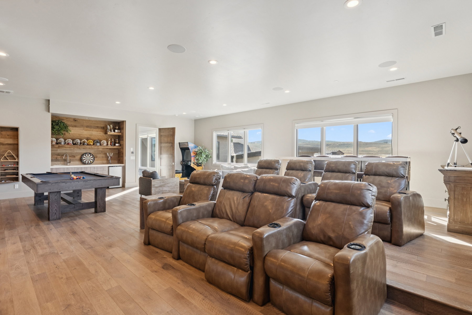 Spacious entertainment area featuring tiered seating, a dedicated wet bar with built-in shelving, large windows, and wood-finish flooring