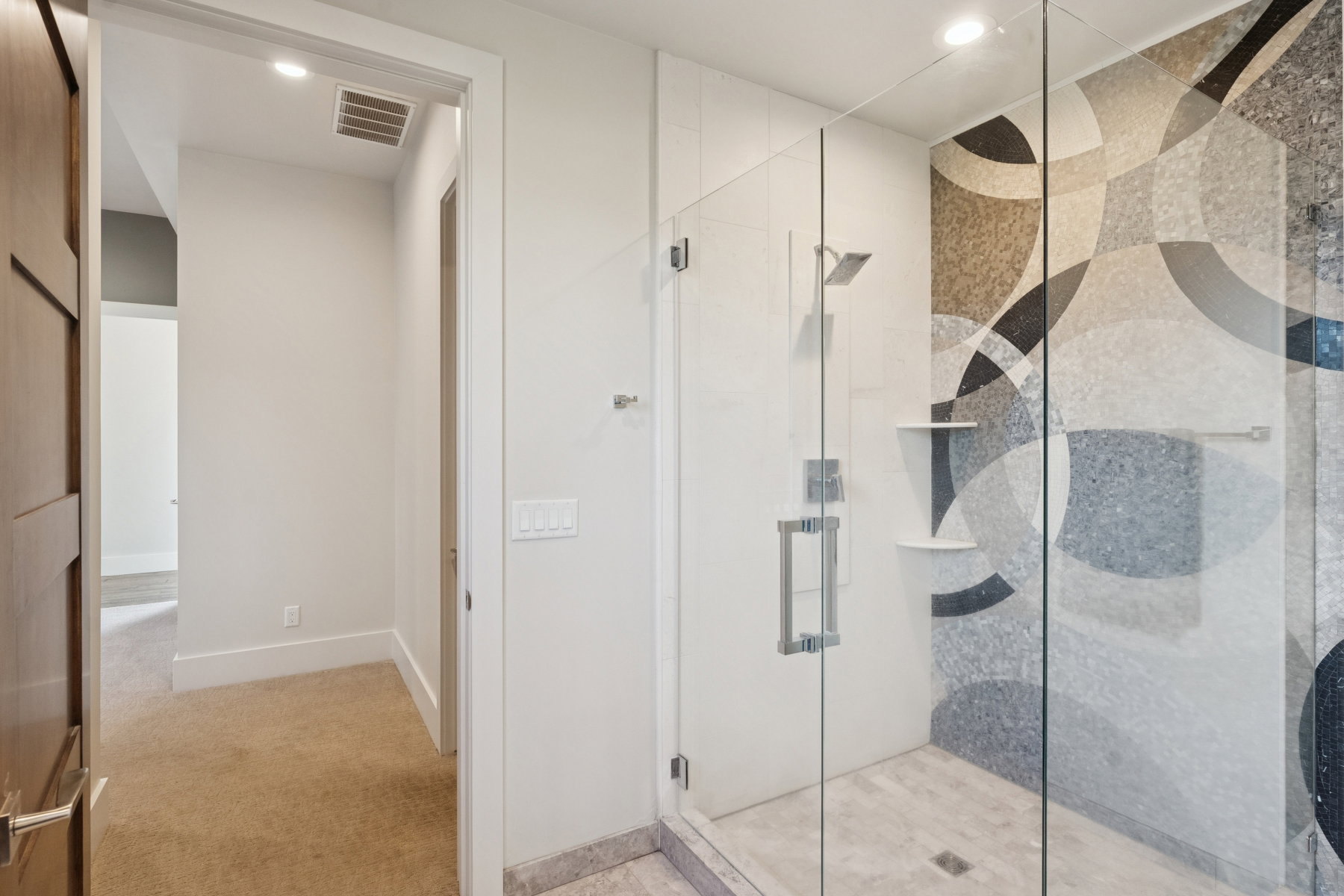 Enclosed glass shower featuring an artistic mosaic tile pattern with geometric shapes, a square rain shower head, and built-in shelving