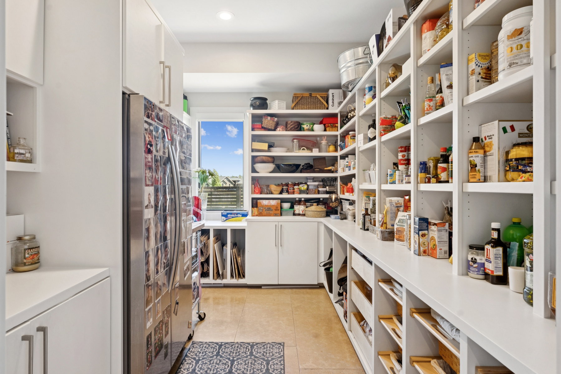 Extensive pantry with ample white shelving, integrated cabinetry, stainless steel refrigerator, stone-look tile flooring, and natural light from a horizontal window