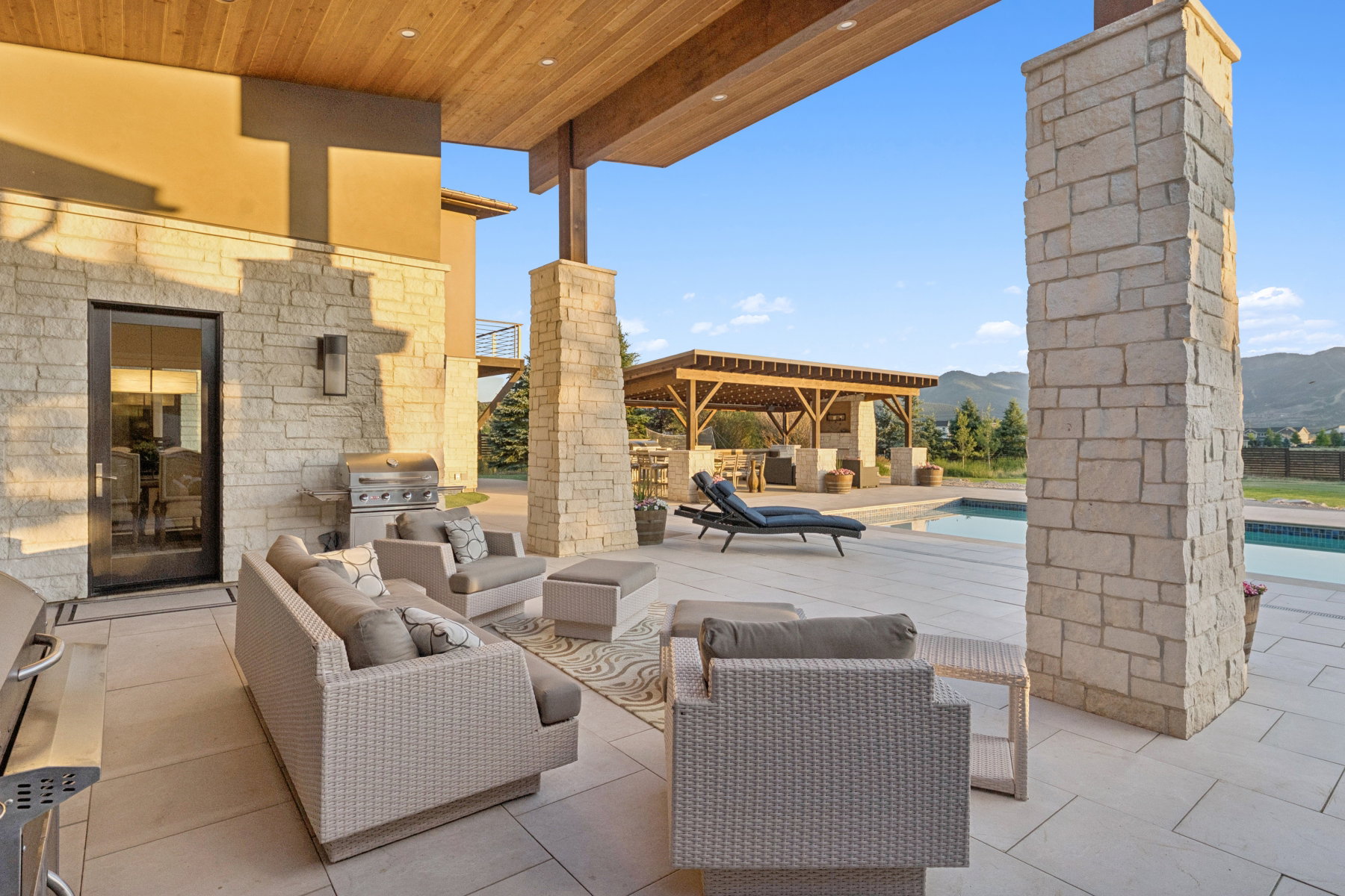 Expansive stone patio featuring a built-in outdoor kitchen area, a swimming pool, and a dedicated pergola structure