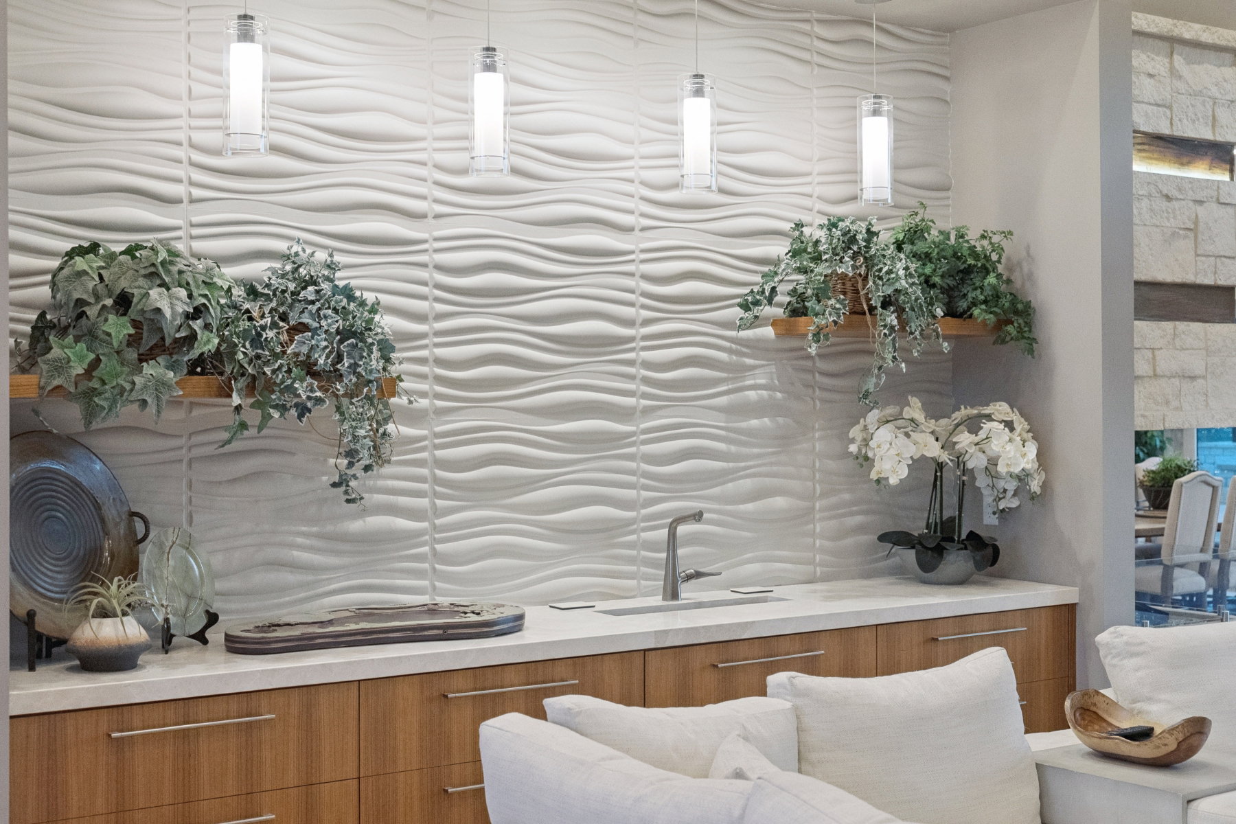 Wet bar featuring a textured wave-pattern accent wall, three pendant lights, wood-finish cabinetry with linear hardware, and a solid surface countertop with an undermount sink and gooseneck faucet