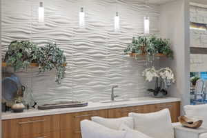 Wet bar featuring a textured wave-pattern accent wall, three pendant lights, wood-finish cabinetry with linear hardware, and a solid surface countertop with an undermount sink and gooseneck faucet