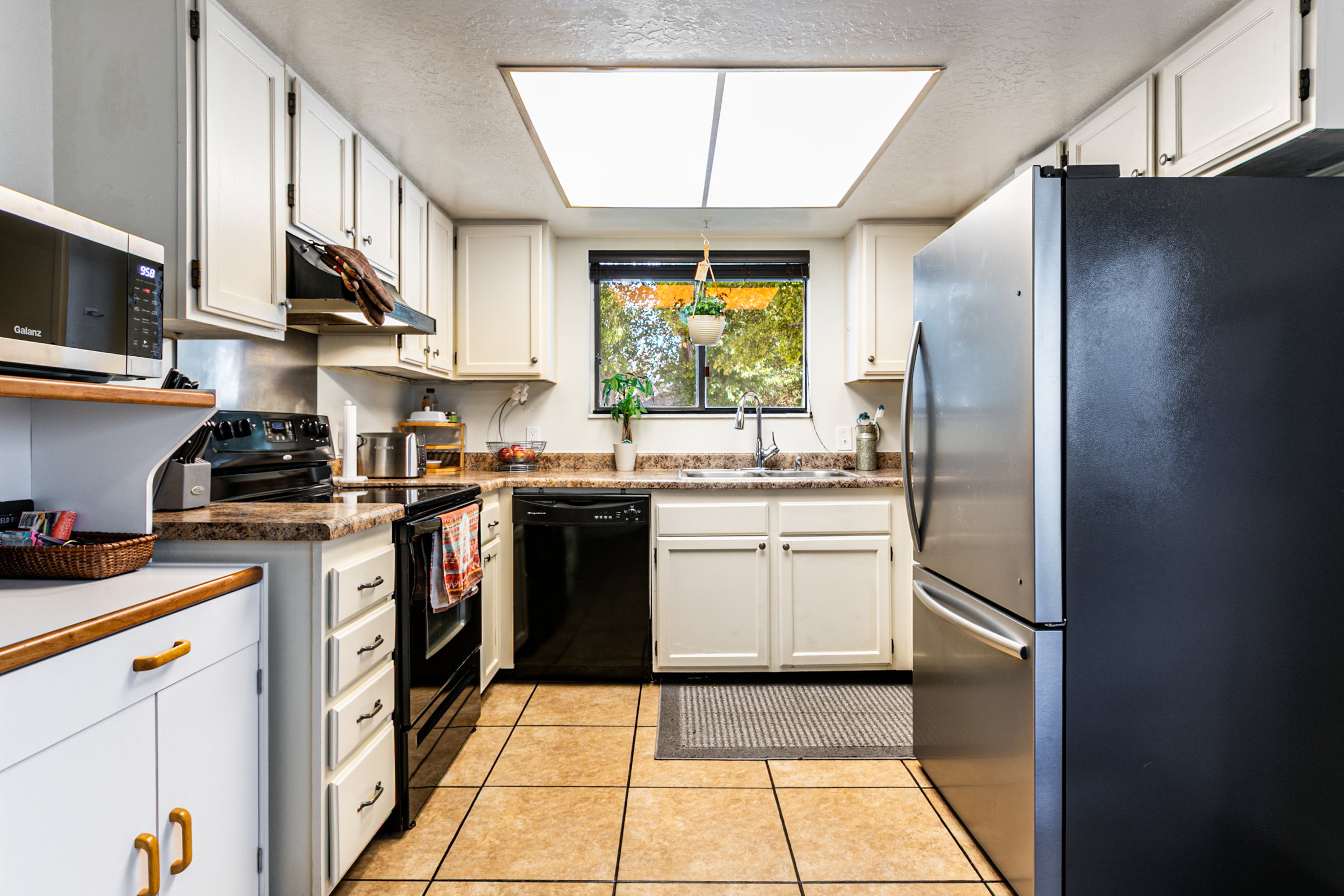 Kitchen featuring white cabinetry, a window over the sink, black appliances, tan tile flooring, and a large overhead light fixture