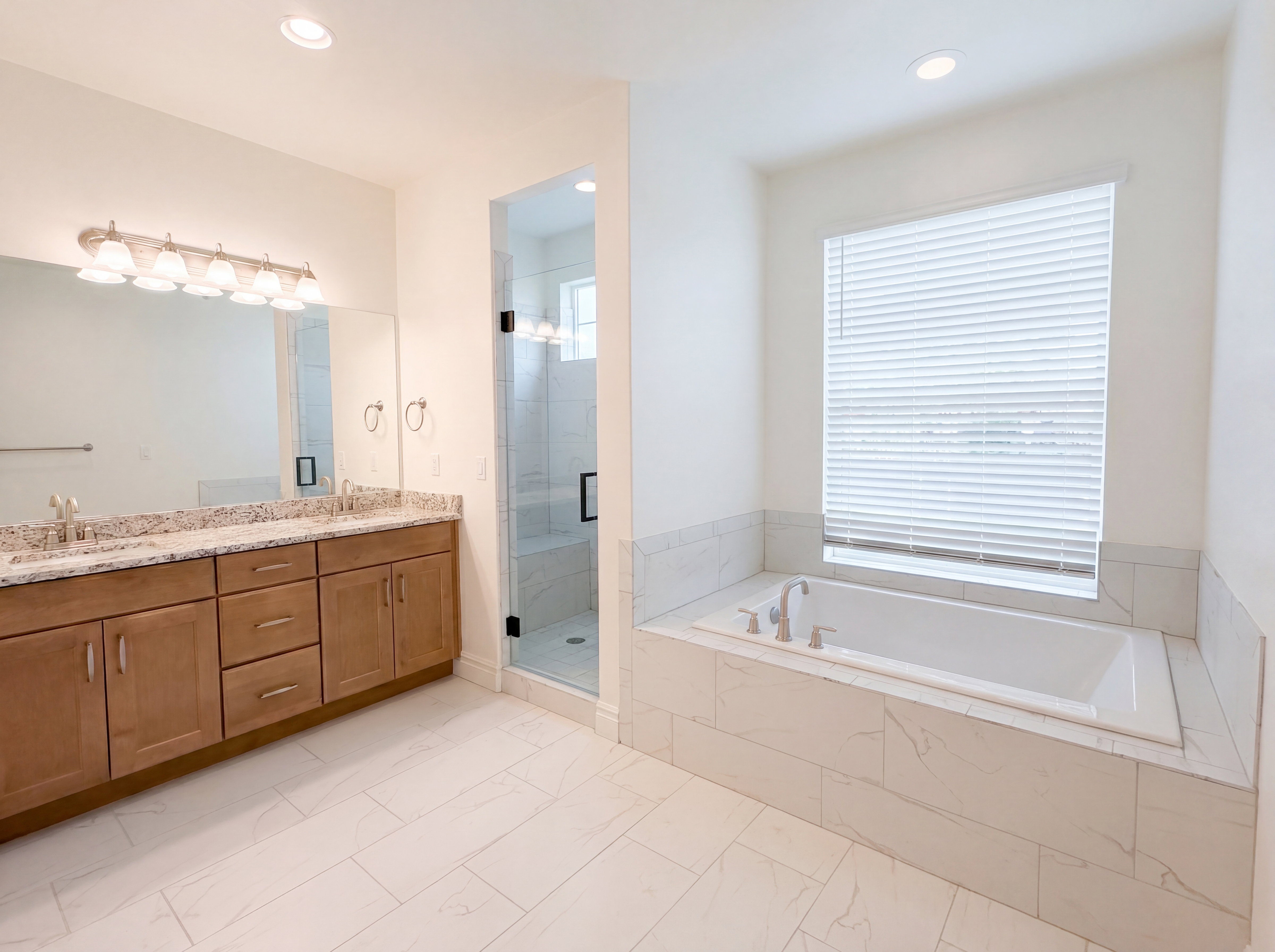 Spacious bathroom featuring a built-in soaking tub, a clear glass shower enclosure, a double vanity with light-toned cabinetry, and extensive tile flooring