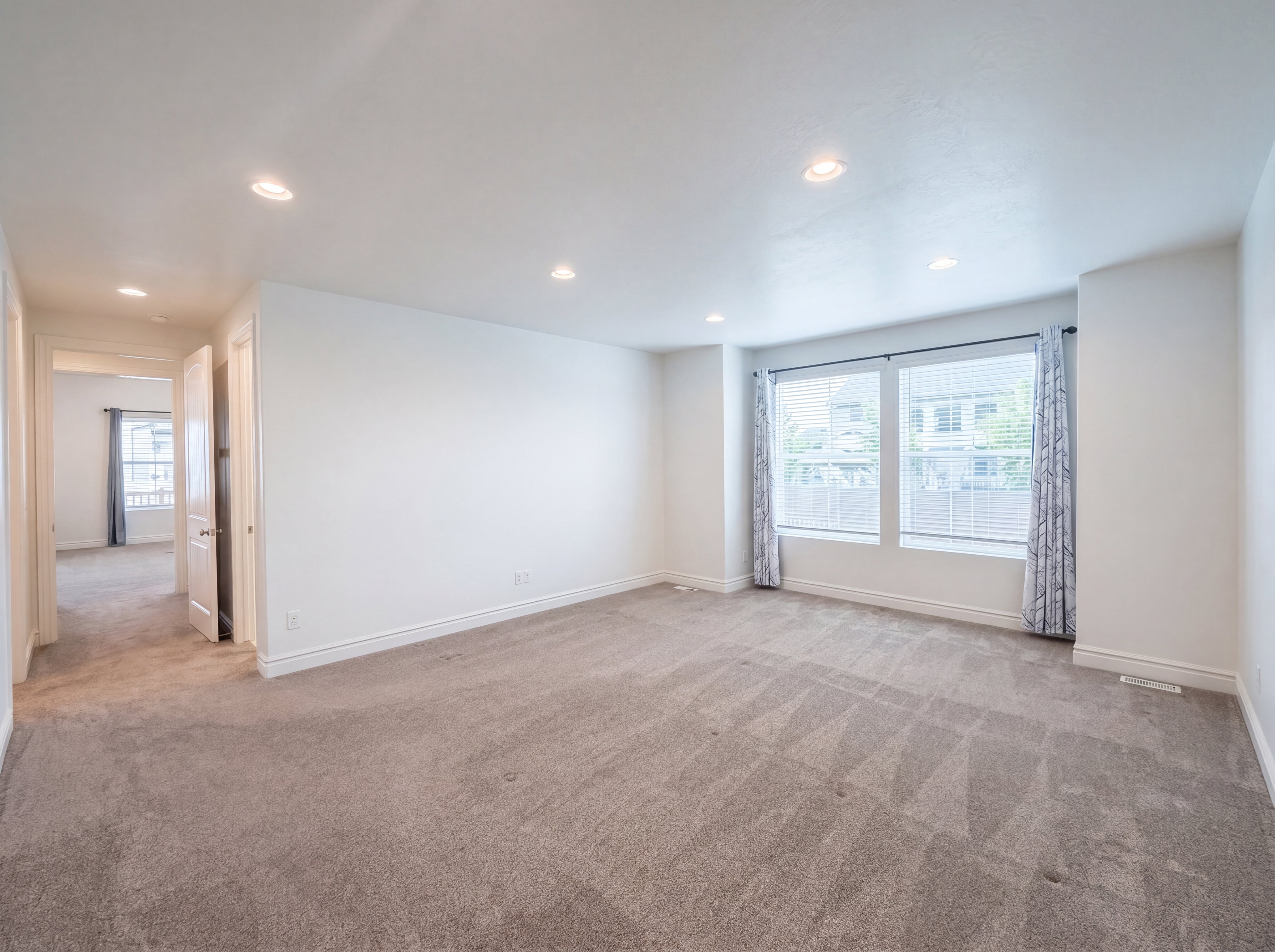 Spacious room featuring neutral carpeting, multiple recessed ceiling lights, and two large windows with blinds and curtains