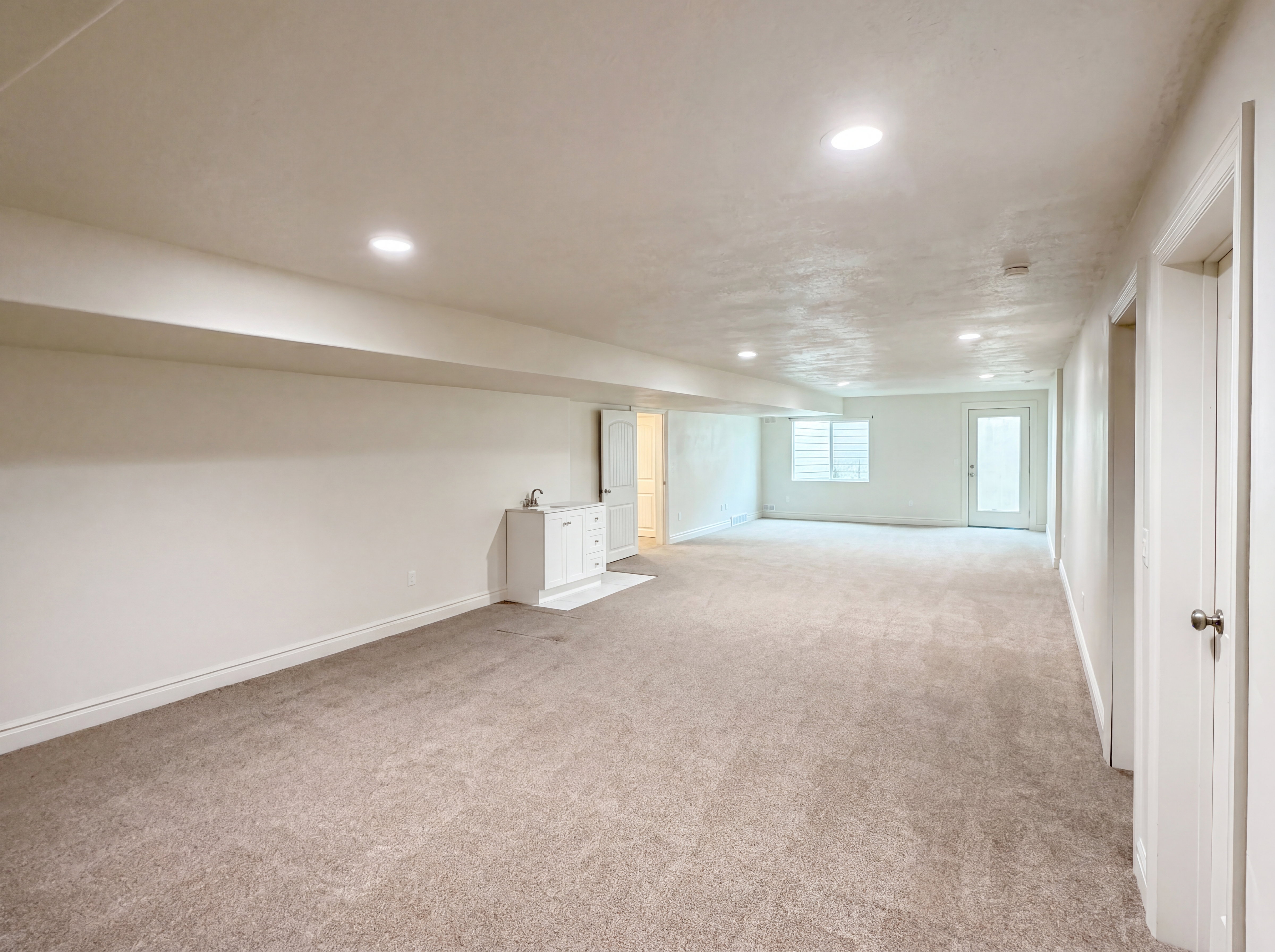 Spacious carpeted room featuring recessed lighting, a built-in wet bar with cabinet storage, a window with horizontal blinds, and a glass-paneled exterior door