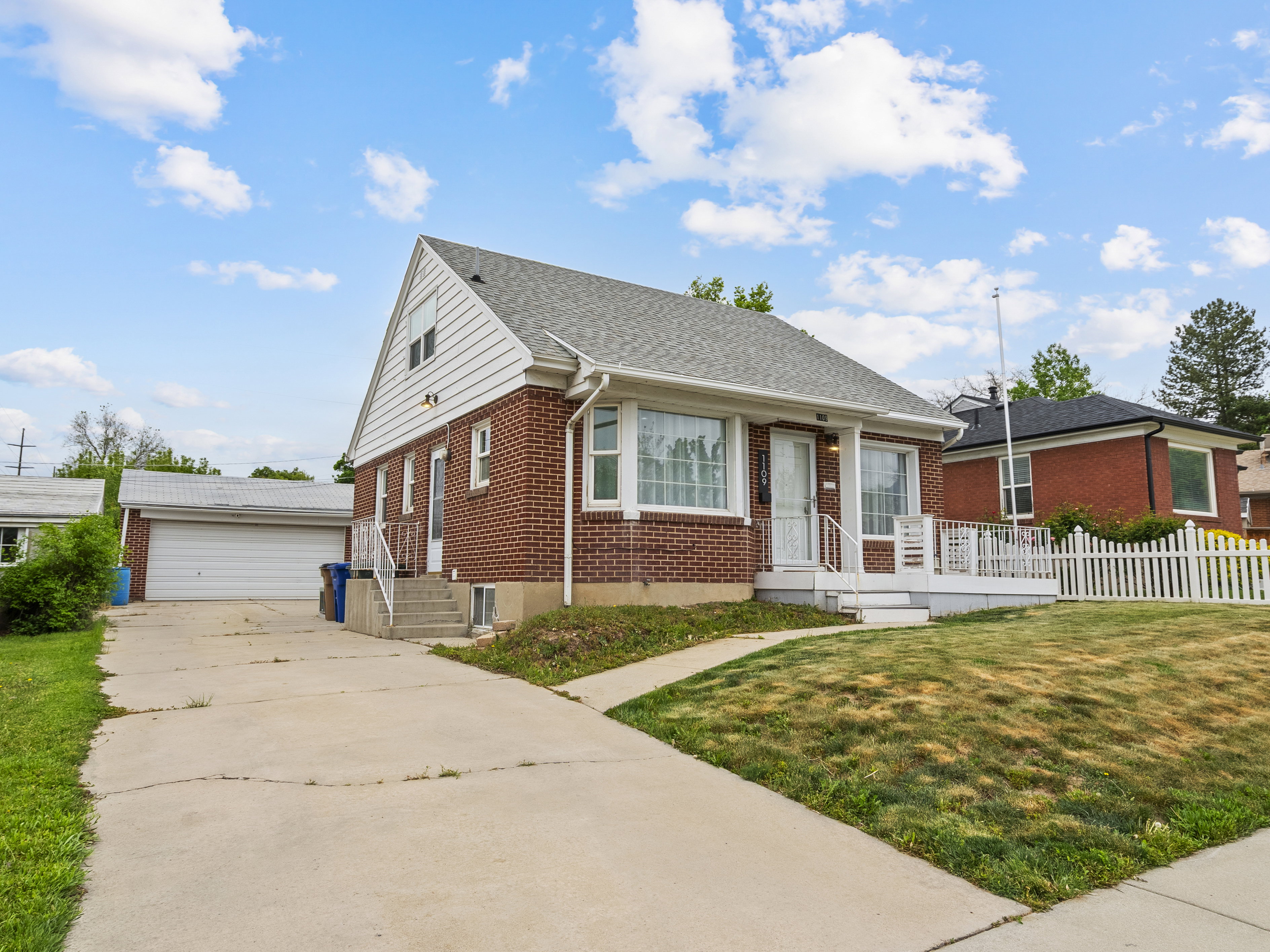 Brick exterior home featuring a white picket fence, covered front porch, and a detached two-car garage with an extended driveway