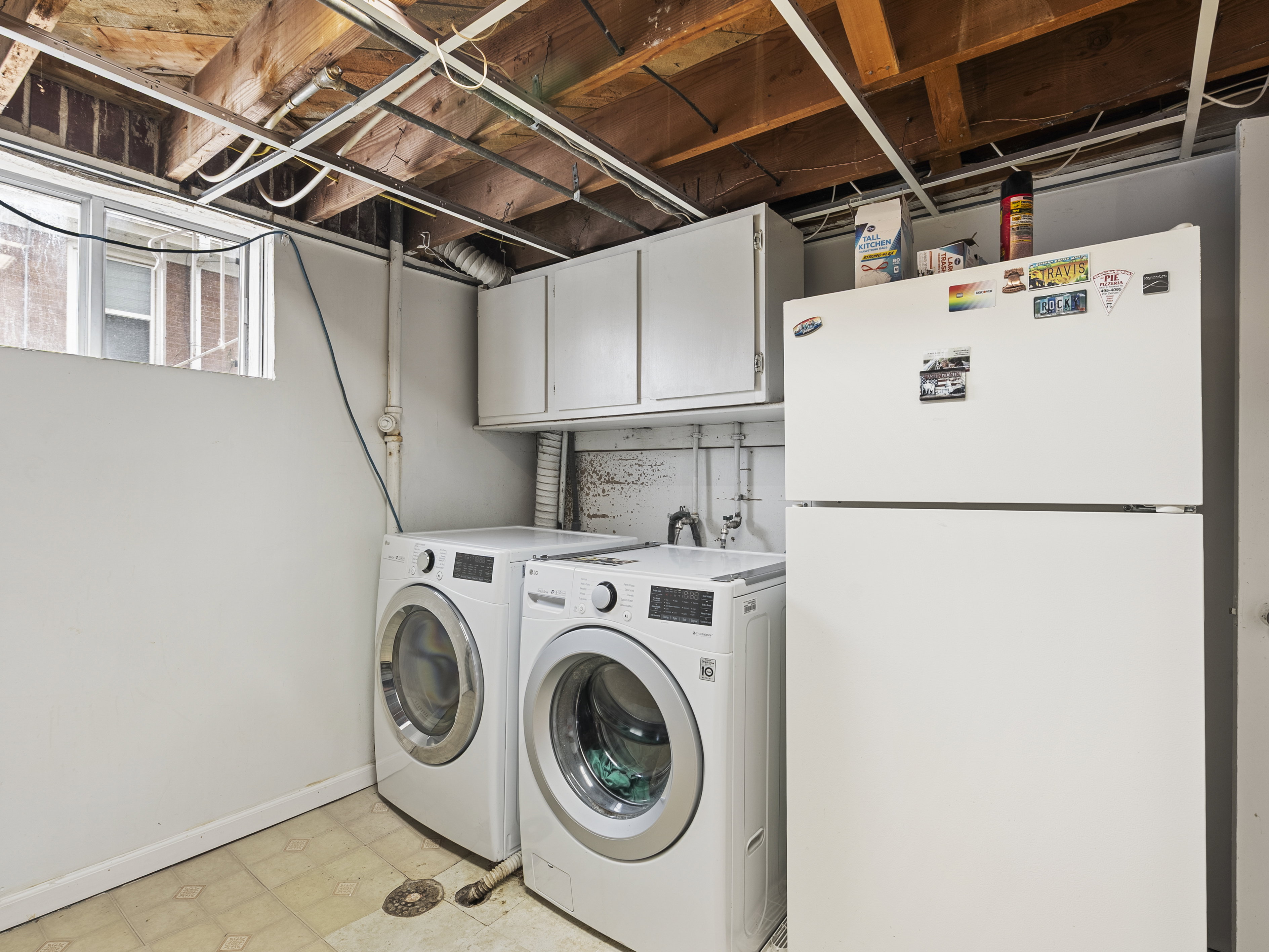 Dedicated laundry area with two front-loading machines, overhead white cabinetry, exposed wood ceiling joists, and a utility sink