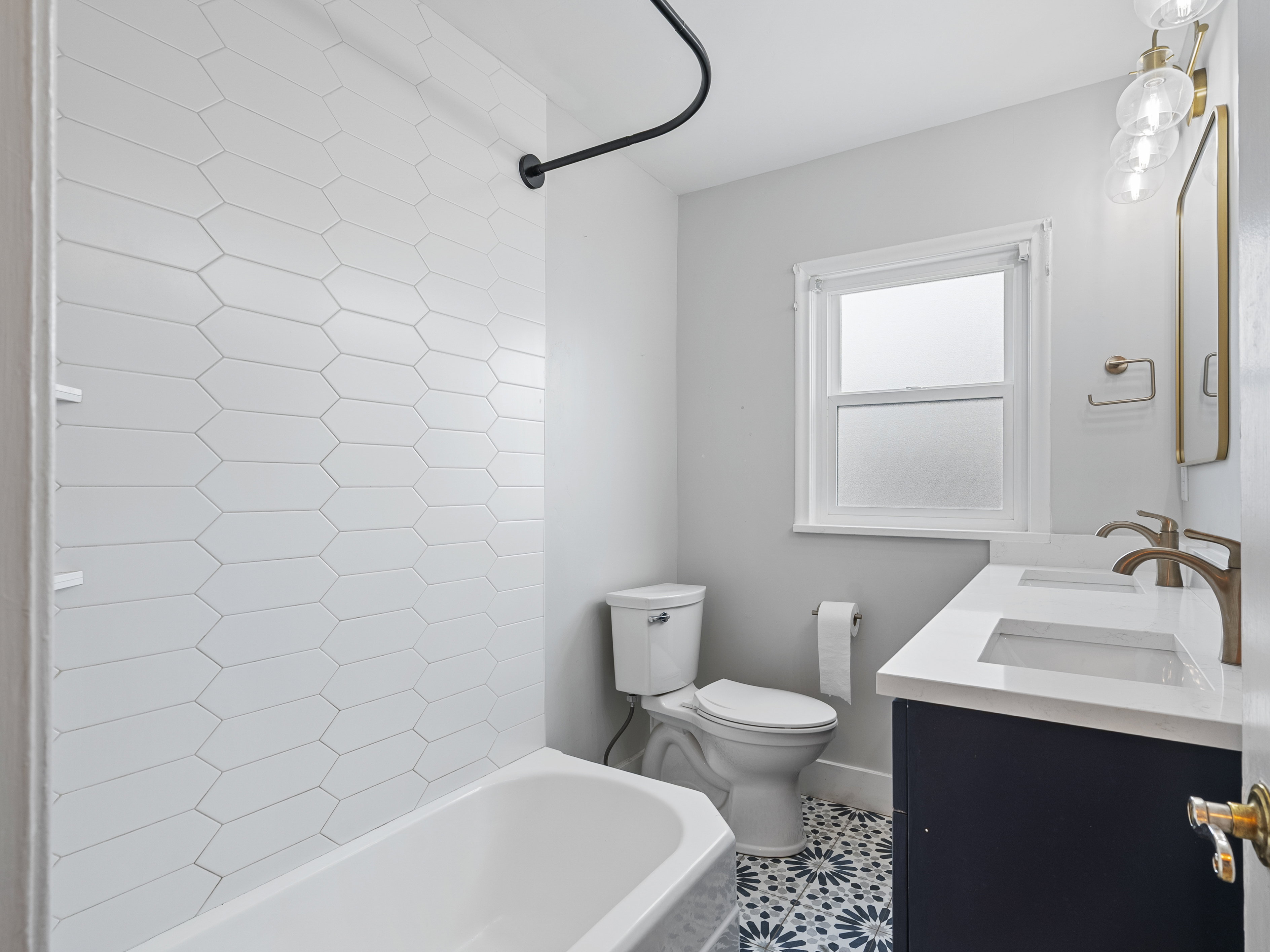 Bathroom featuring a white subway-tiled shower, black shower rod, window with frosted glass, dual vanity with white countertop, and patterned floor tiling