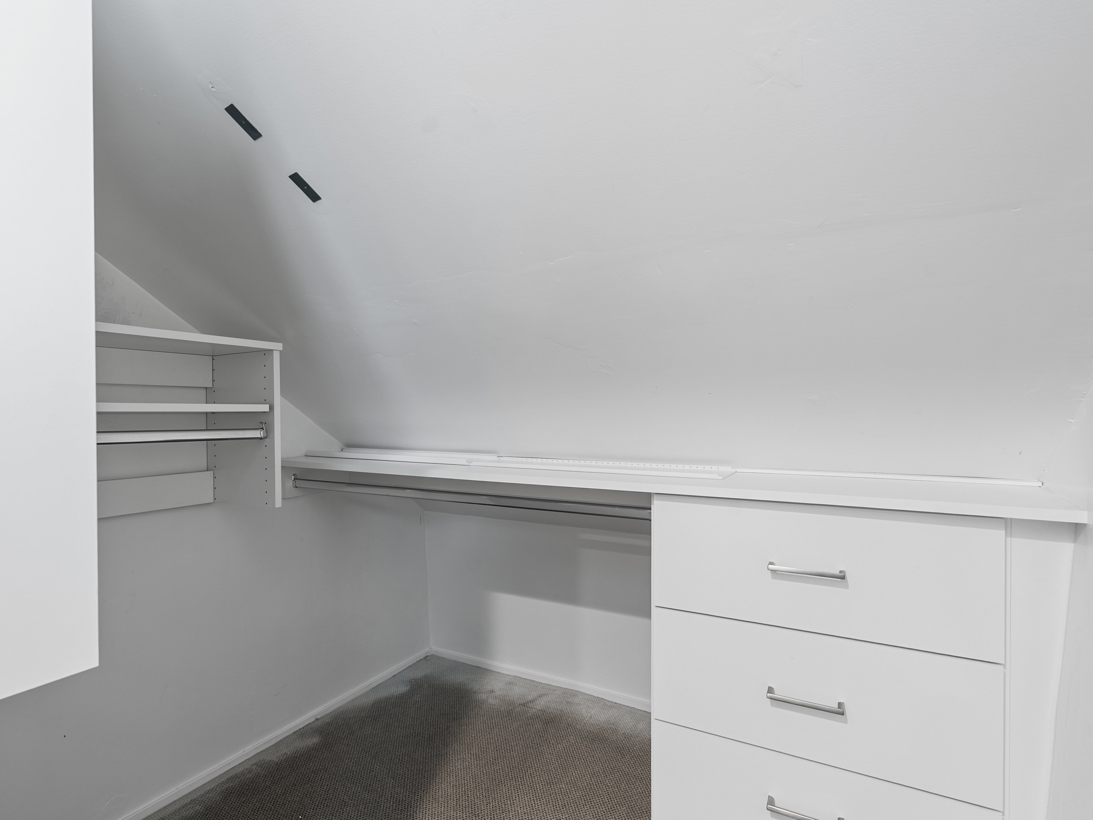 Sloped ceiling closet featuring built-in shelving, hanging rods, and a three-drawer dresser with chrome pulls