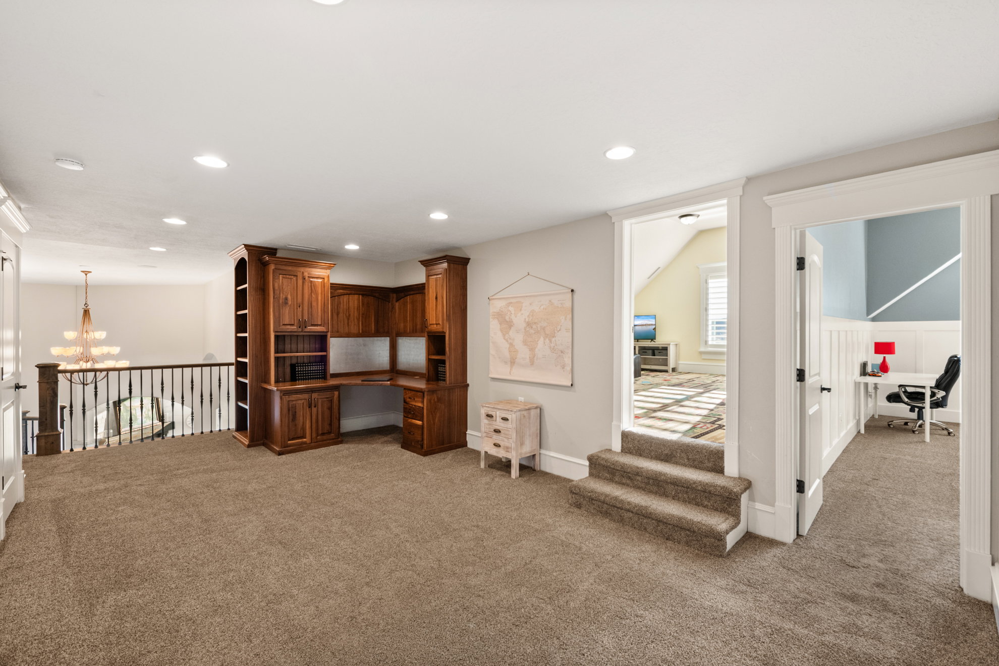 Expansive carpeted loft area featuring a built-in wood-finish desk with integrated shelving, recessed lighting, and a wrought iron banister overlooking the floor below