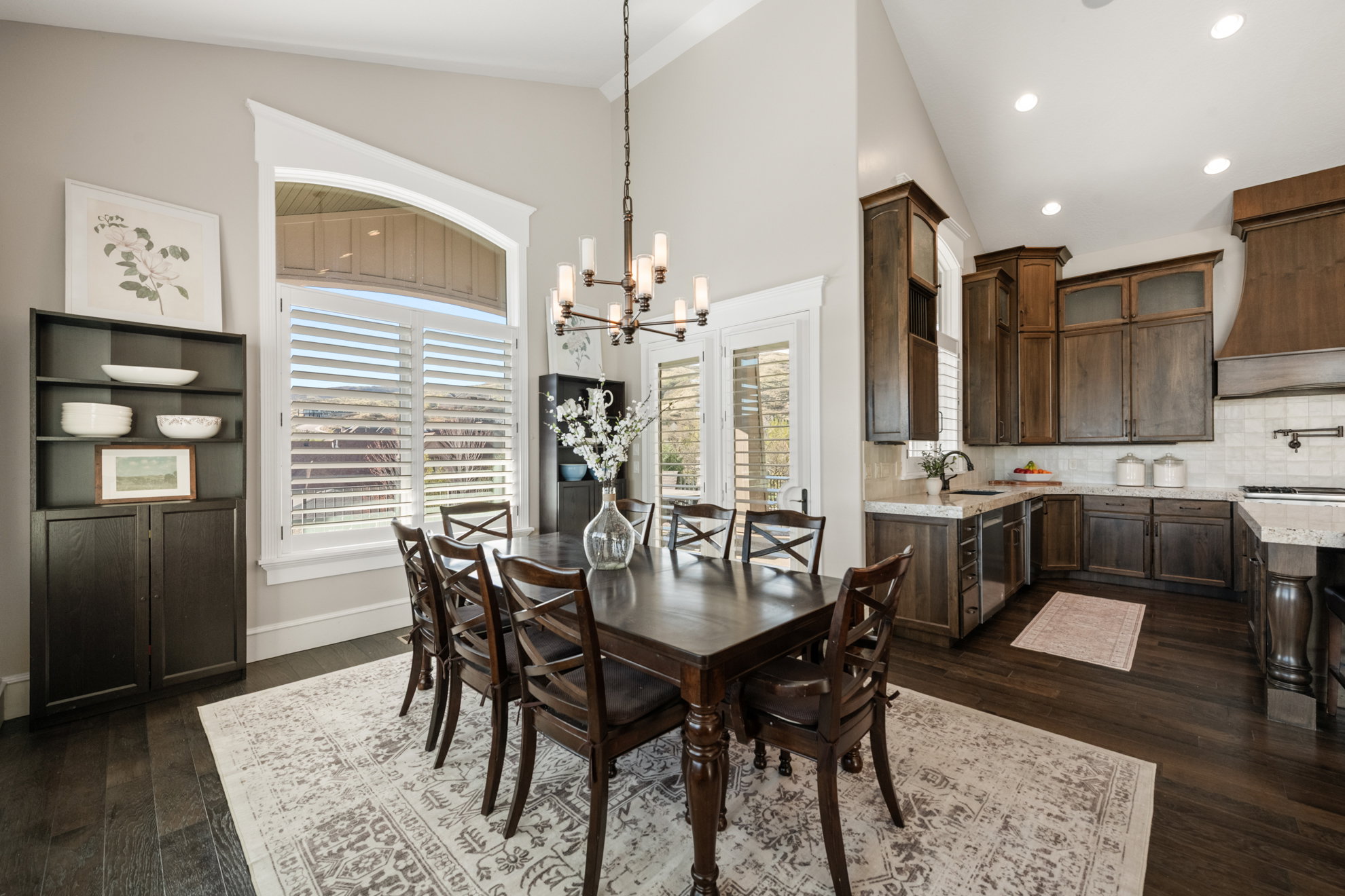 Open-concept dining area featuring a vaulted ceiling with a chandelier, dark wood-finish flooring, and white plantation shutters