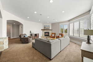 Spacious living area featuring a stone-clad fireplace with a wood mantel, a series of large windows with white plantation shutters, an arched doorway, recessed lighting, and a neutral carpet