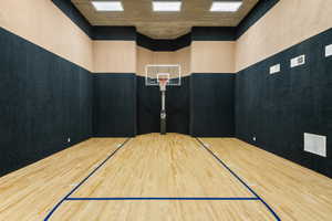 Indoor court featuring wood-finish flooring with blue demarcation lines, a basketball hoop with a glass backboard, sound-dampening wall panels, and recessed ceiling lighting