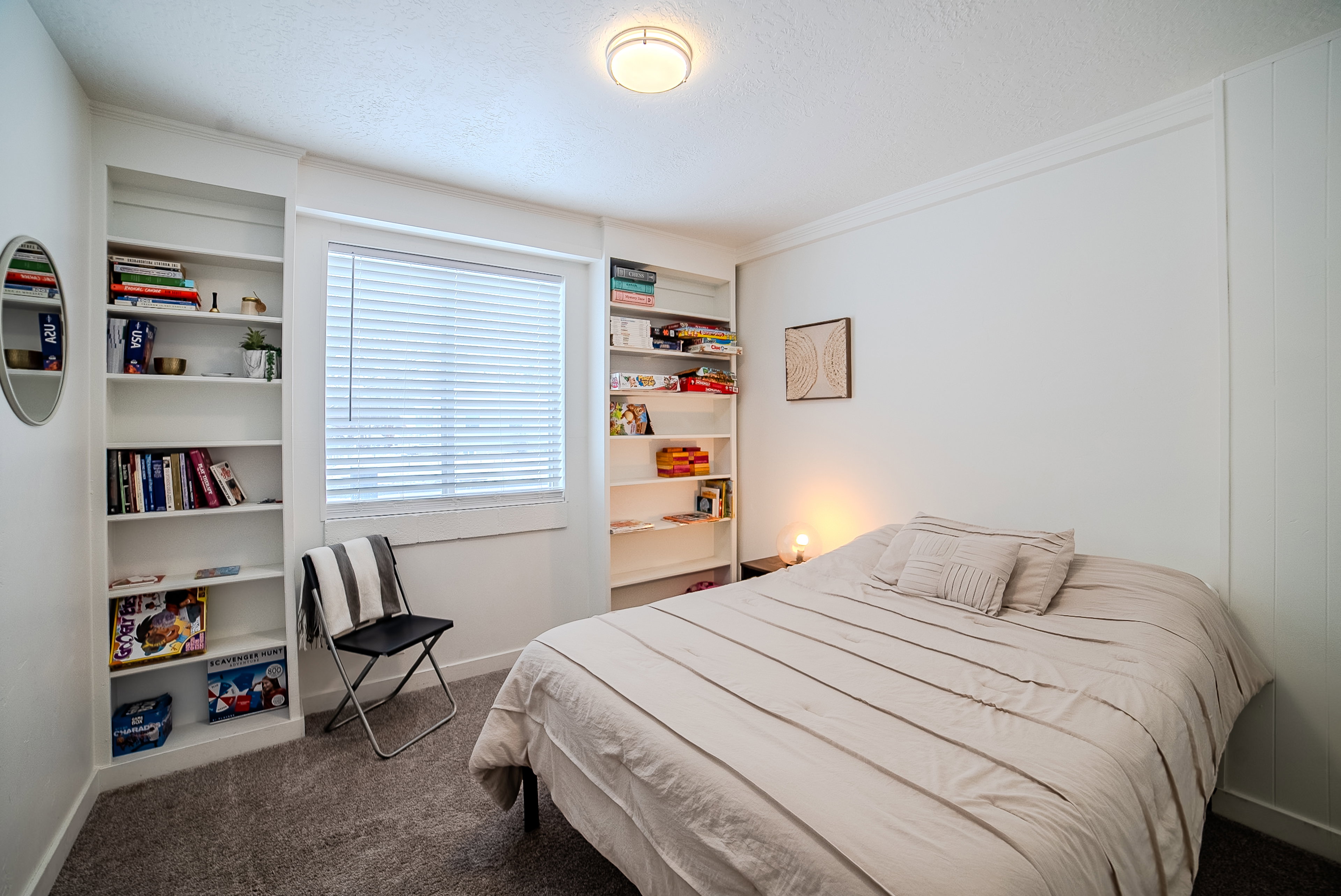 Bedroom featuring a window with blinds, two built-in shelving units, wall paneling, and textured carpeting