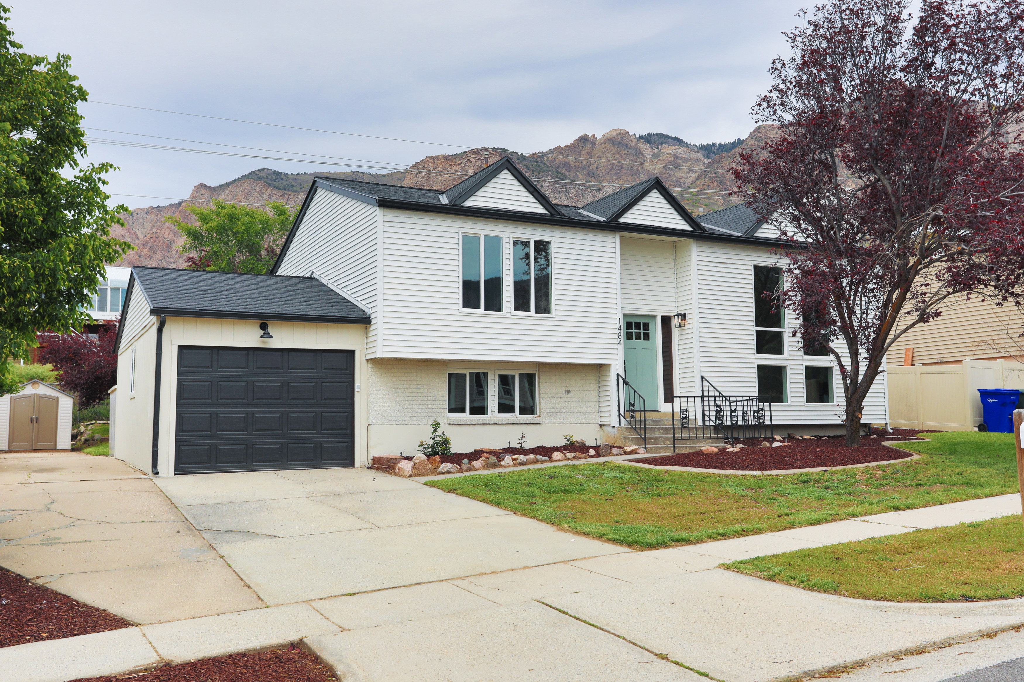 White siding exterior with a dark shingle roof, featuring a two-car garage, established front yard landscaping, a light blue front door, and a concrete driveway