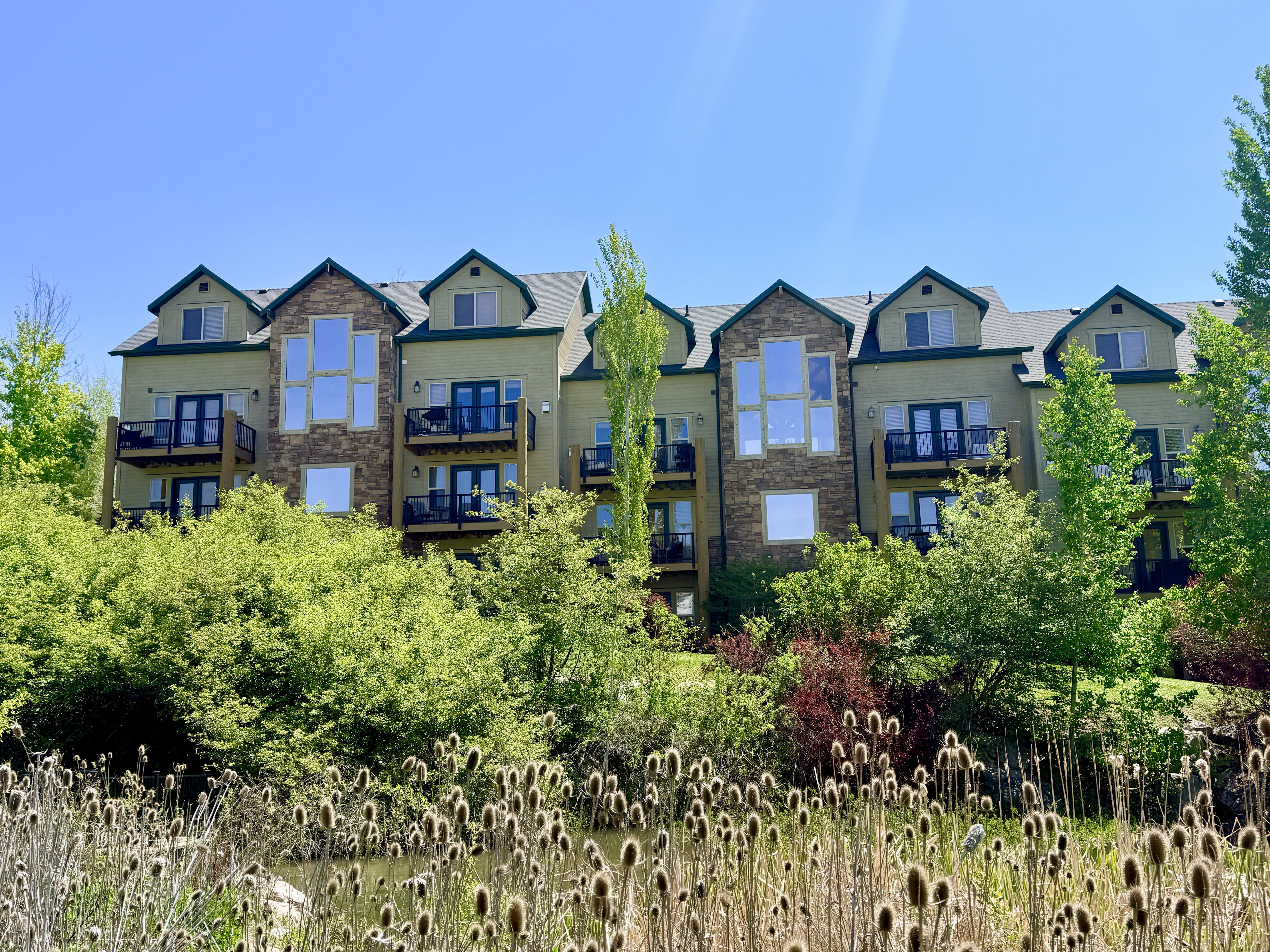Multi-story building featuring stone accents, multiple private balconies with black railings, dormer windows, and lush surrounding greenery