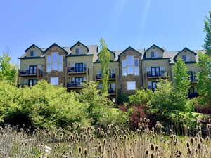 Multi-story building featuring stone accents, multiple private balconies with black railings, dormer windows, and lush surrounding greenery