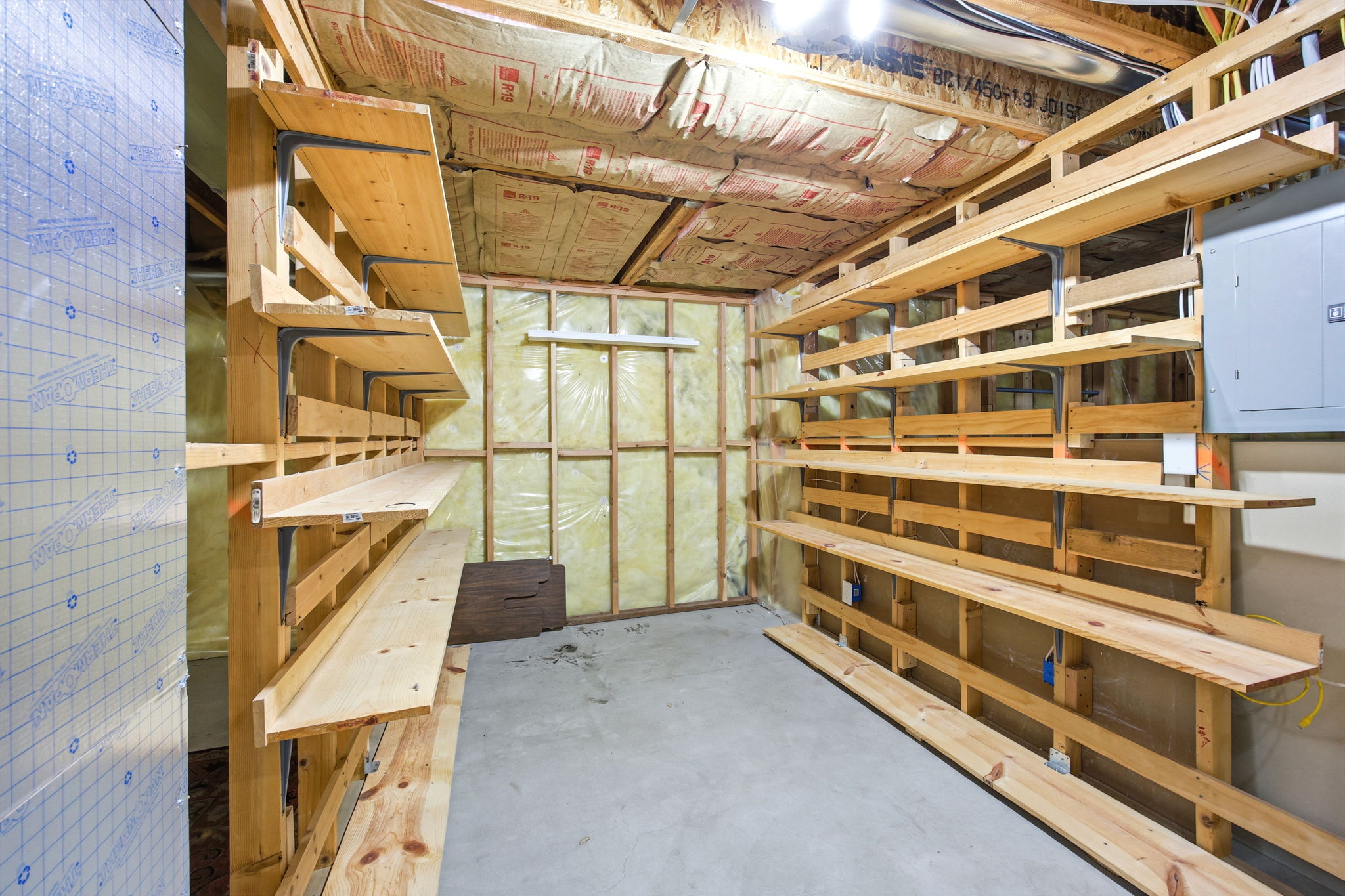 Utility room featuring extensive wood shelving, concrete flooring, exposed insulation, and a visible electrical panel