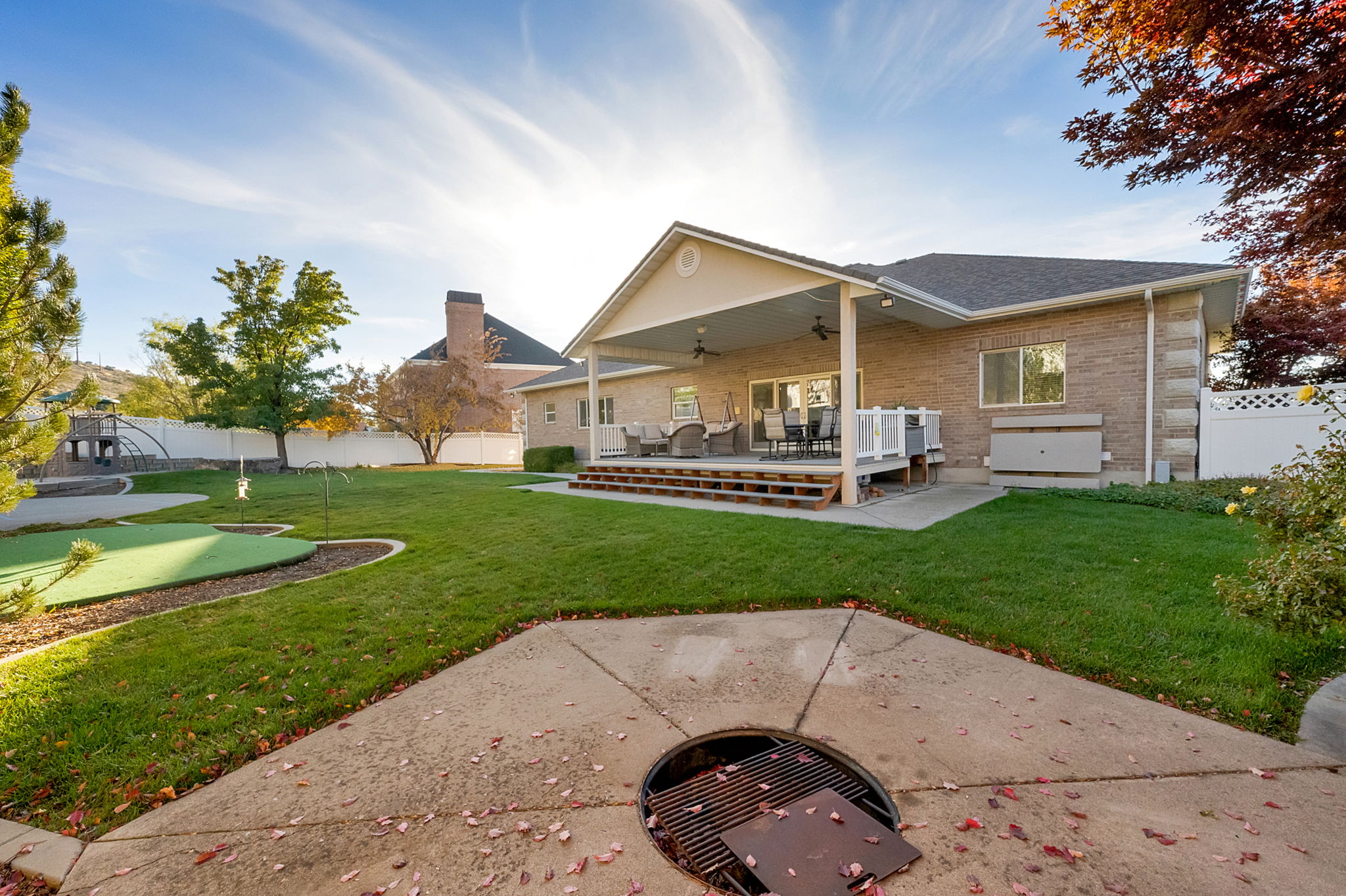 Expansive backyard featuring a covered deck, brick exterior, putting green, mature landscaping, and a built-in fire pit