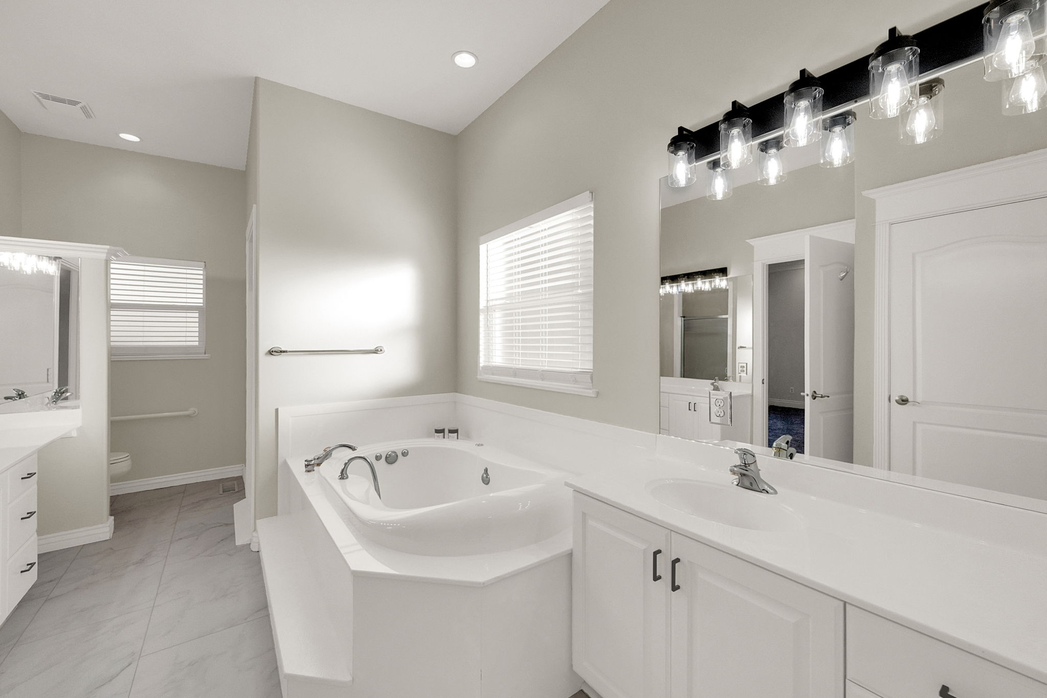 Spacious bathroom featuring a built-in jetted tub, dual vanities with white cabinetry, light countertops, and large format tile flooring
