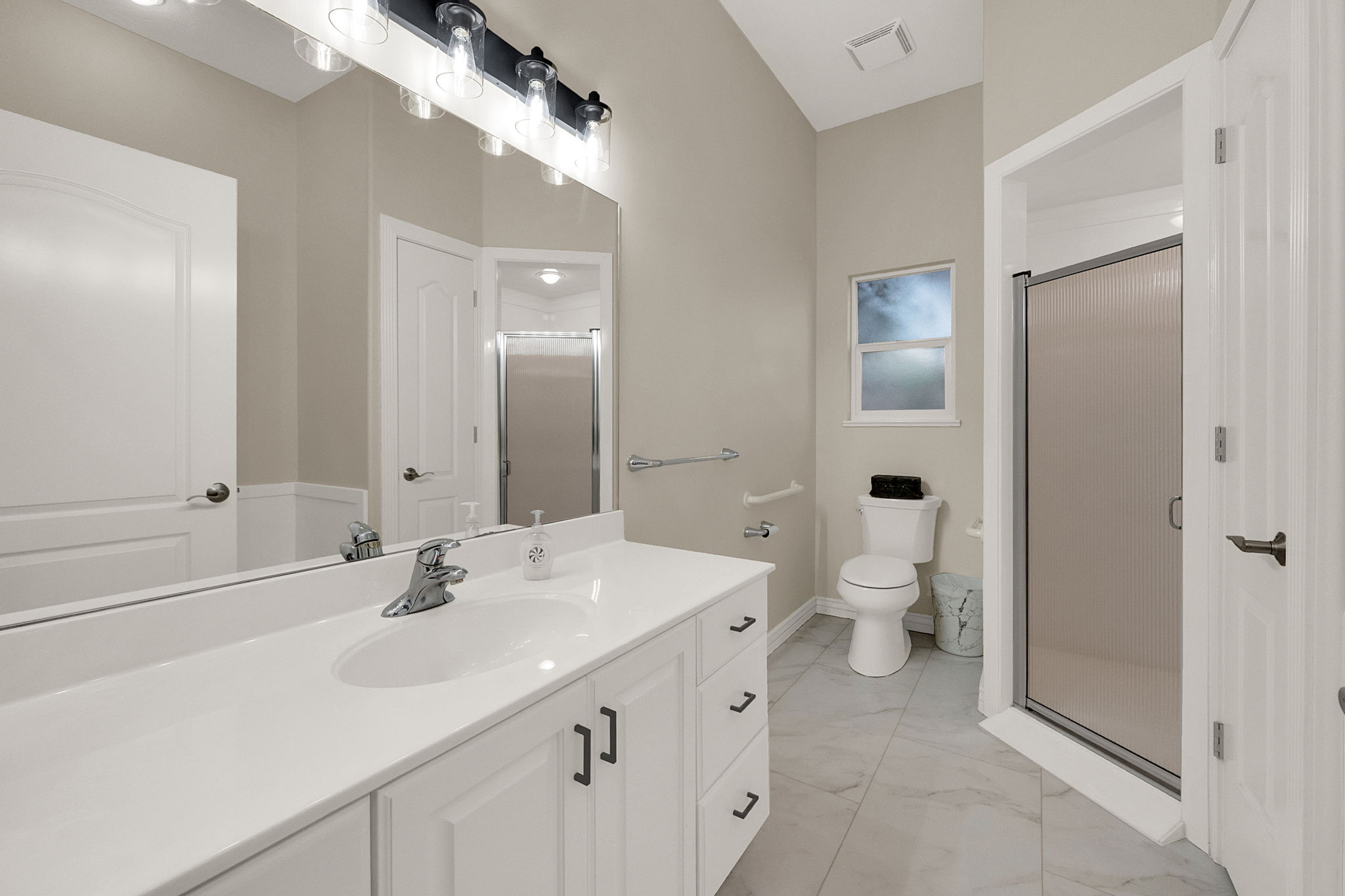 Spacious bathroom featuring a white vanity with integrated sink, large mirror, black and clear glass vanity lighting, tiled flooring, and a glass-enclosed shower
