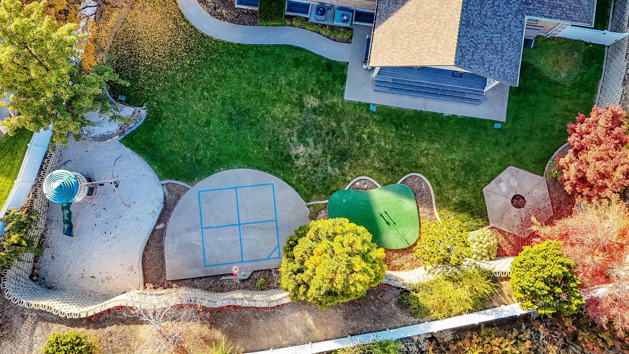 Expansive backyard featuring a lush lawn, mature trees, concrete patio with a four-square court, dedicated sand play area with a slide structure, and a putting green