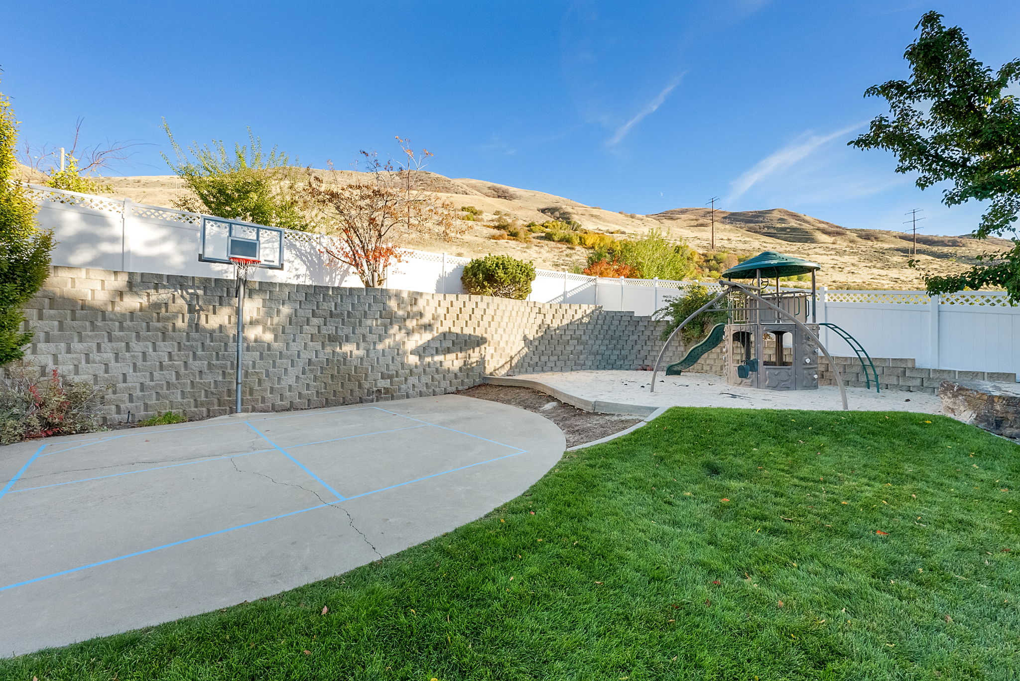 Expansive backyard featuring a concrete sport court with a basketball hoop, a sand play area, a multi-level playset with a slide, and a manicured lawn