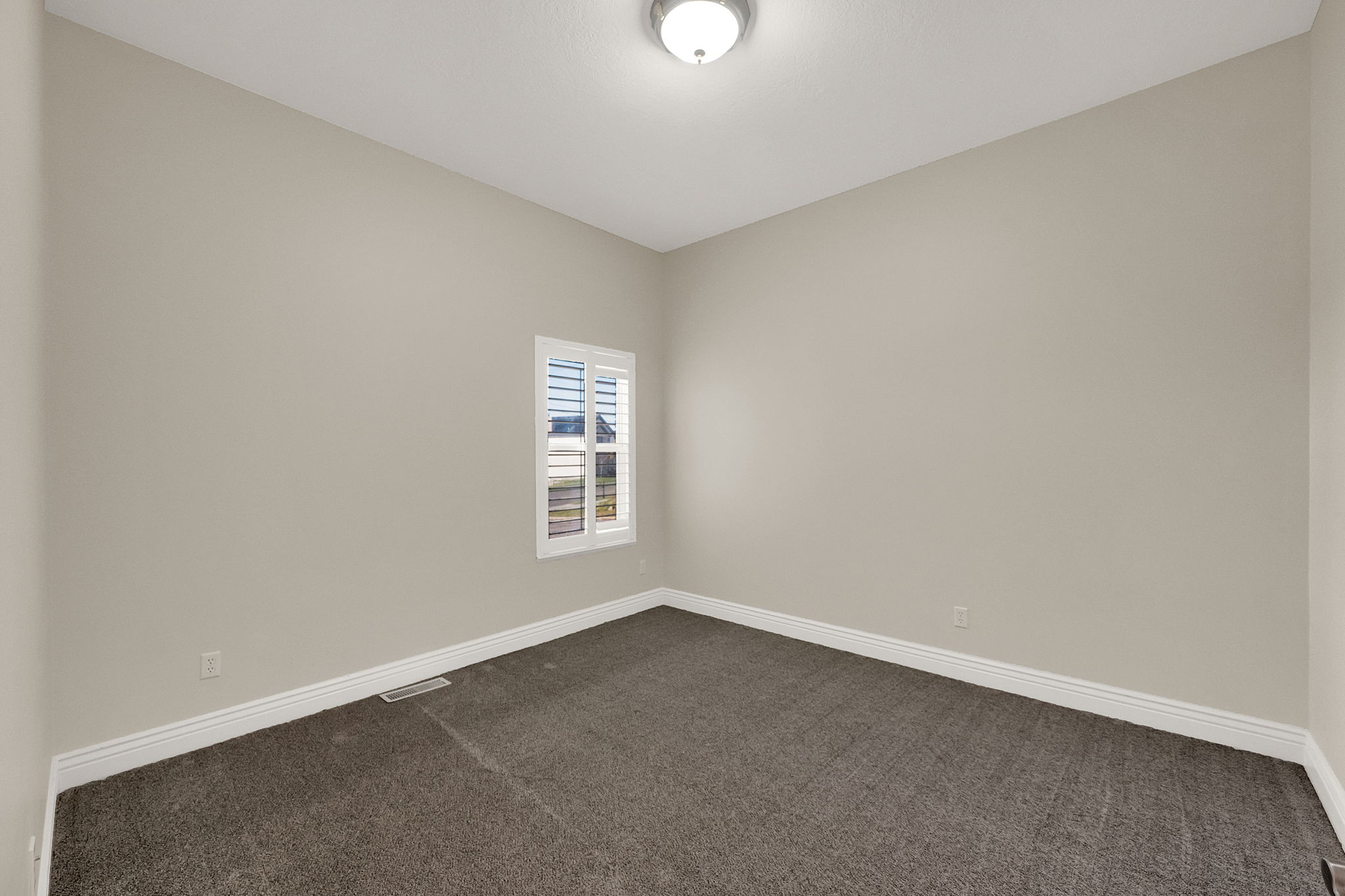 Neutral-toned room featuring a single window with white plantation shutters, neutral carpet flooring, and a flush-mount ceiling light fixture