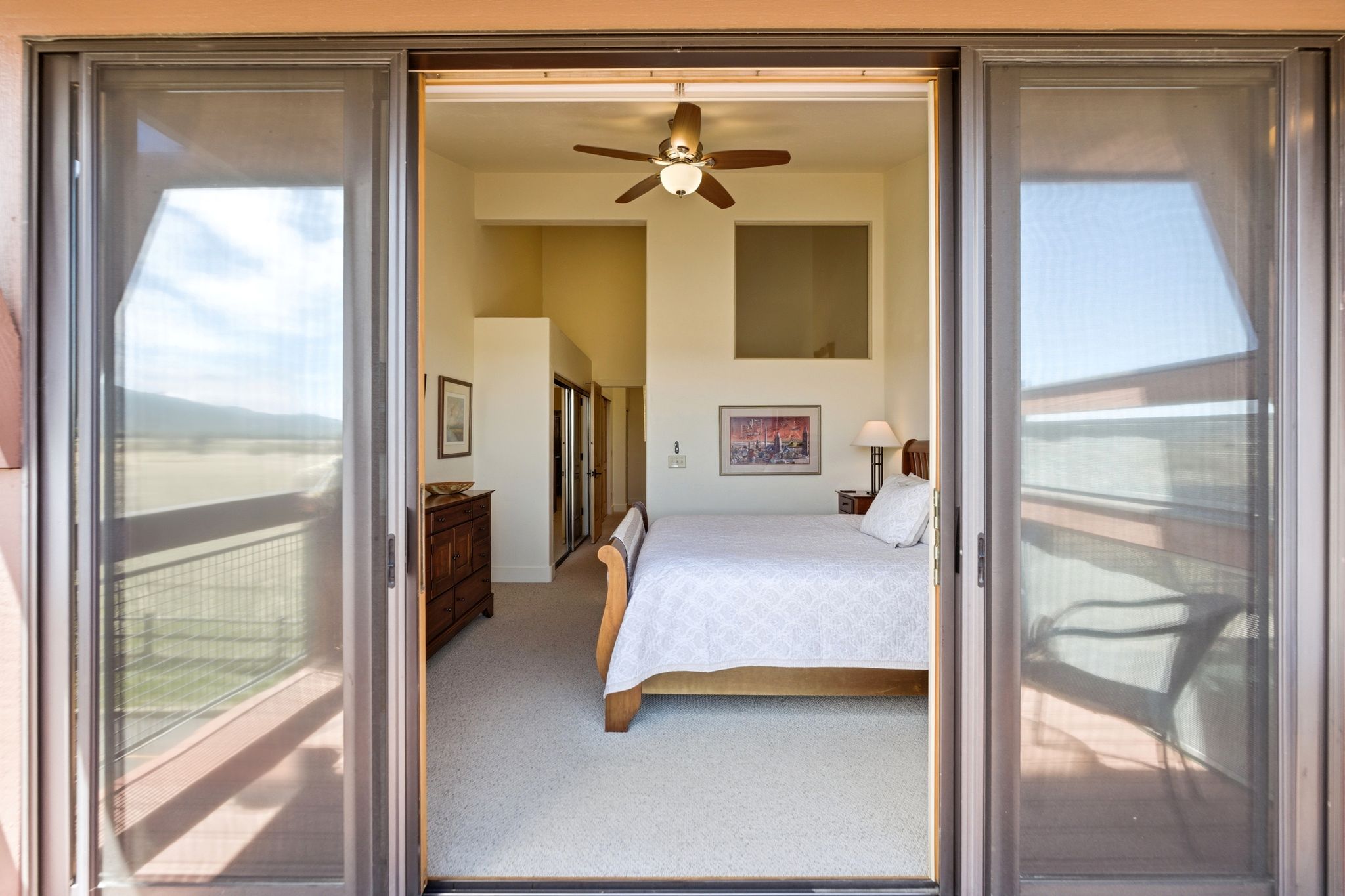 Sliding glass doors opening to a bedroom featuring a vaulted ceiling with a ceiling fan, light beige carpet, and a window opening to another interior space