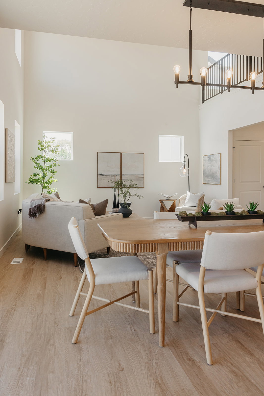 Spacious living area featuring high ceilings, wood-finish flooring, multiple windows, an open staircase with a black metal railing, and an exposed beam chandelier