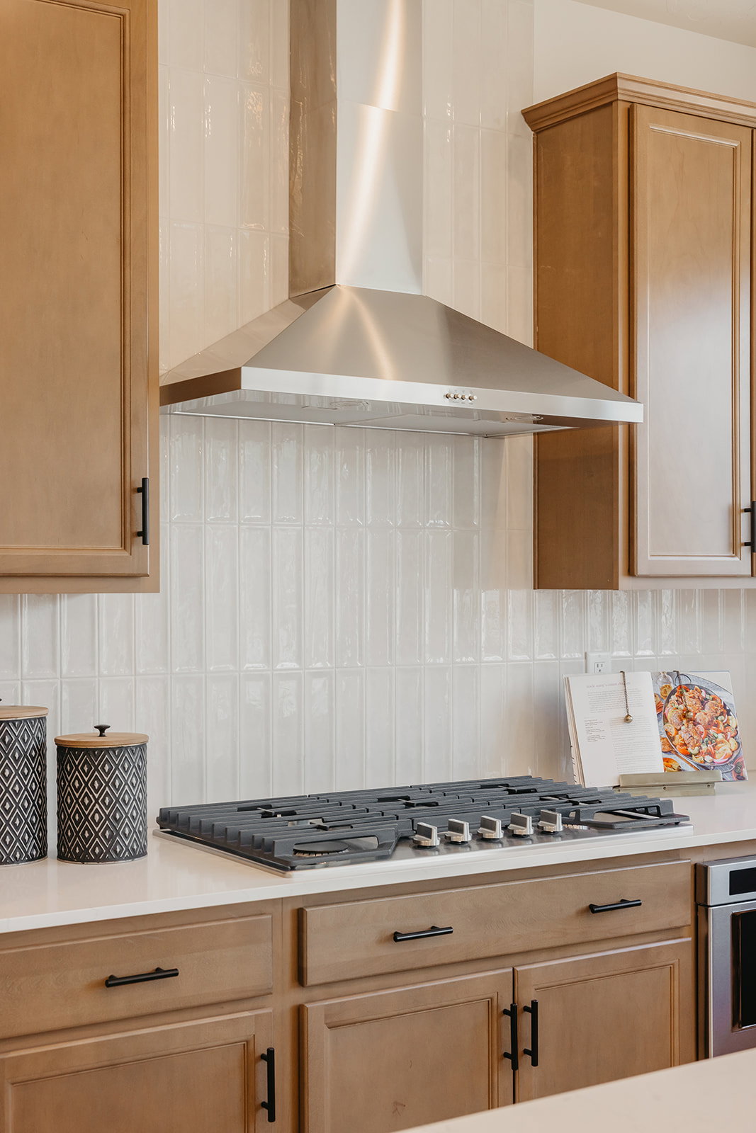 Kitchen featuring a stainless steel range hood, full tile backsplash with vertical rectangular tiles, light wood-finish cabinetry with black hardware, white countertop, and a built-in gas cooktop