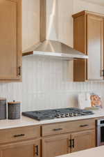 Kitchen featuring a stainless steel range hood, full tile backsplash with vertical rectangular tiles, light wood-finish cabinetry with black hardware, white countertop, and a built-in gas cooktop