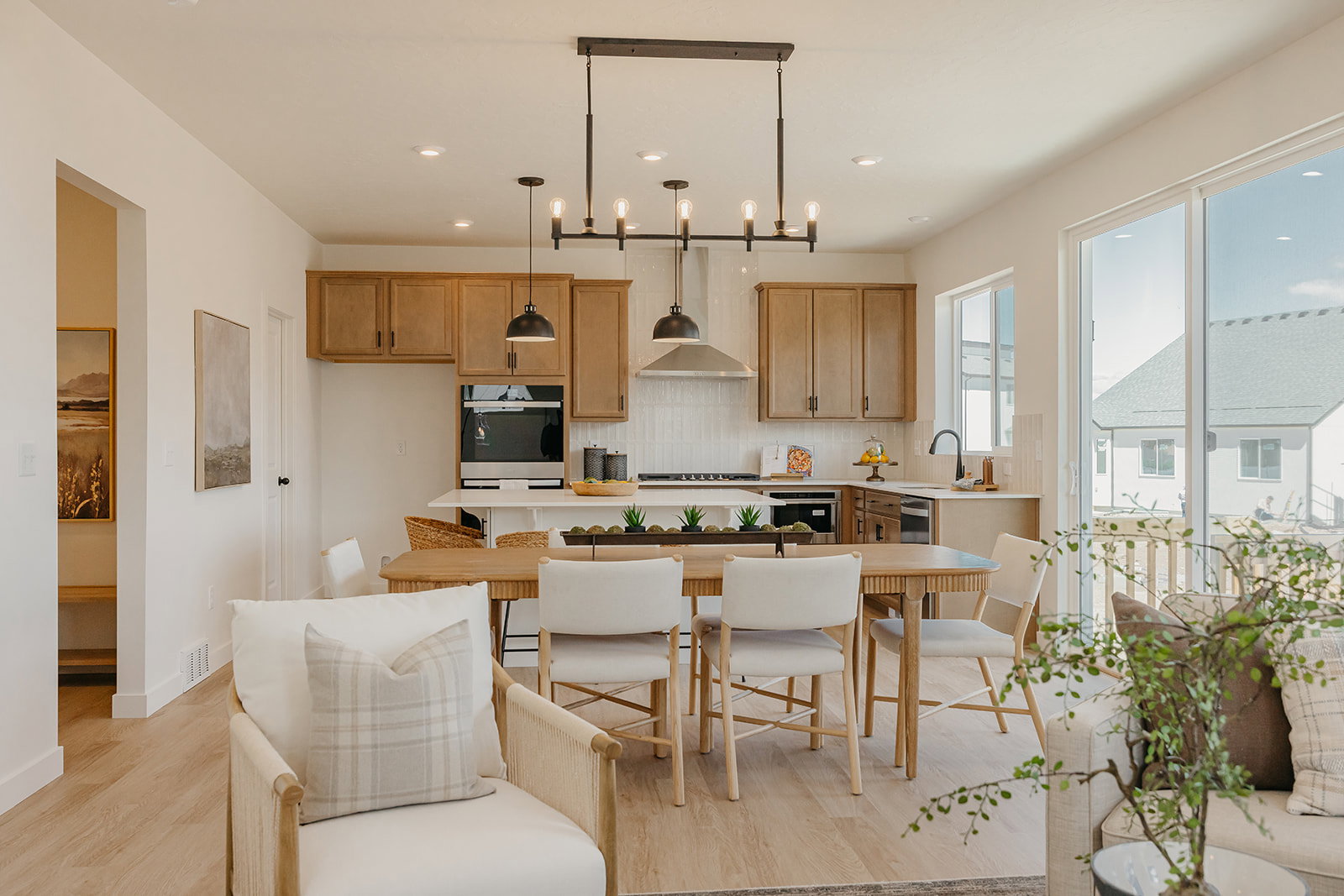 Open-concept living area featuring wood-finish flooring, a contemporary kitchen with light wood cabinetry and light countertops, and a dining space with a linear chandelier