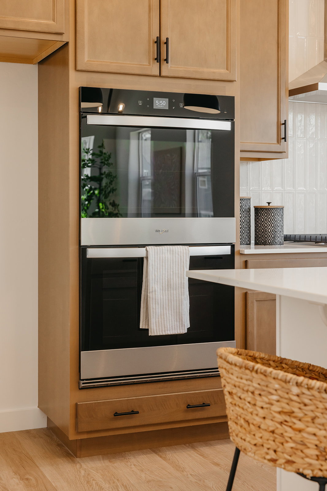 Built-in double oven with stainless steel accents, light wood-finish cabinetry, matte black hardware, white tile backsplash, and light wood-finish flooring