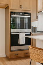 Built-in double oven with stainless steel accents, light wood-finish cabinetry, matte black hardware, white tile backsplash, and light wood-finish flooring