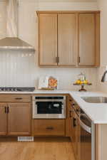 Kitchen featuring light wood-finish cabinetry, white subway tile backsplash, stainless steel range hood, stainless steel built-in microwave, and light countertops with an undermount sink