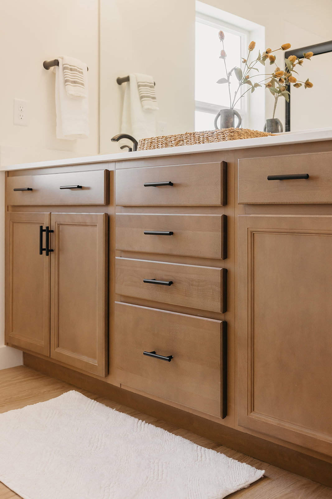 Warm wood-finish cabinetry featuring black hardware, a light-colored countertop, and a large mirror