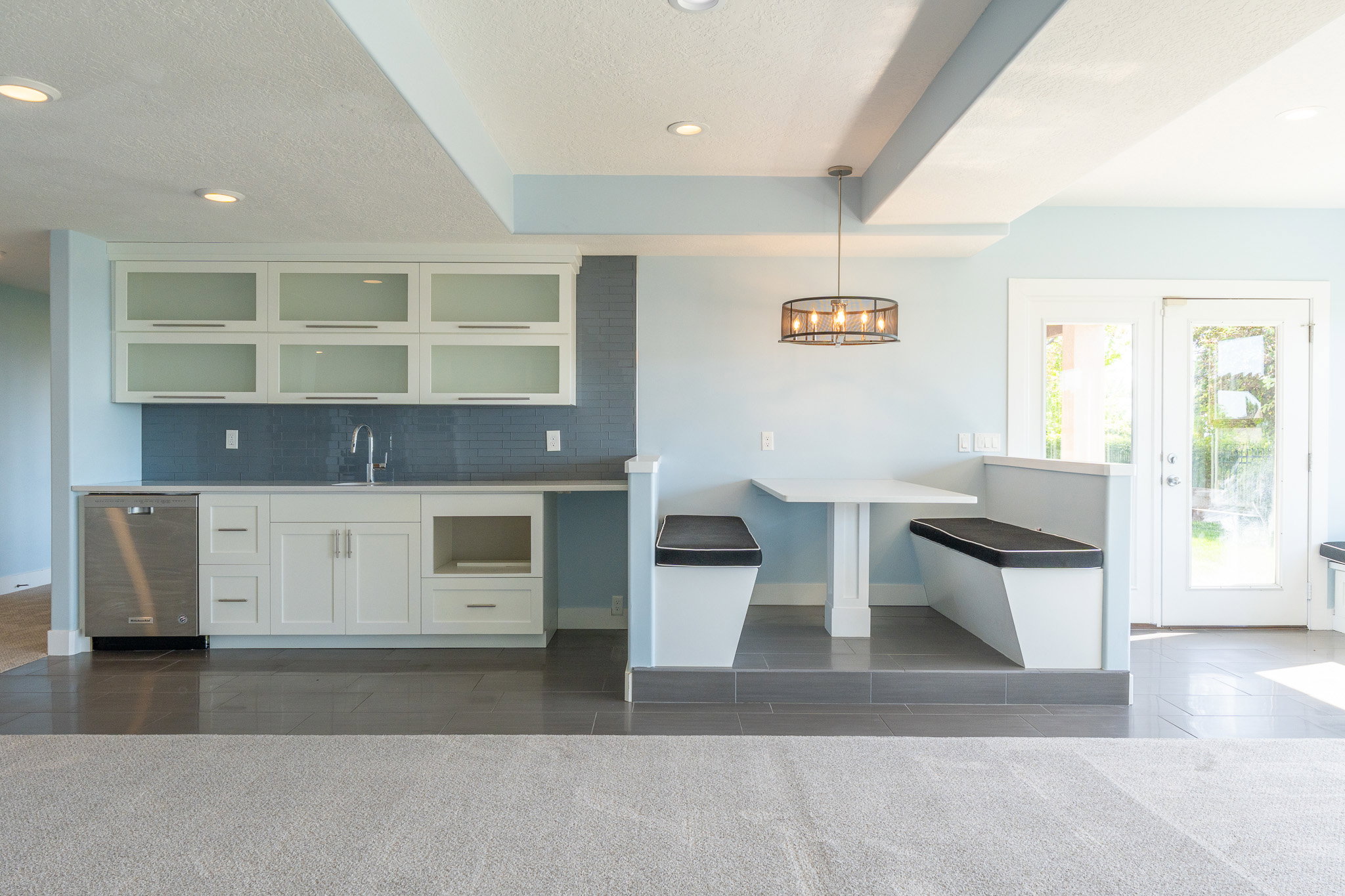 Integrated wet bar featuring white cabinetry with frosted glass uppers, stainless steel dishwasher, and a tile backsplash, alongside a built-in banquette dining area with overhead lighting, and double glass-paned doors