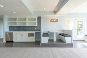 Integrated wet bar featuring white cabinetry with frosted glass uppers, stainless steel dishwasher, and a tile backsplash, alongside a built-in banquette dining area with overhead lighting, and double glass-paned doors