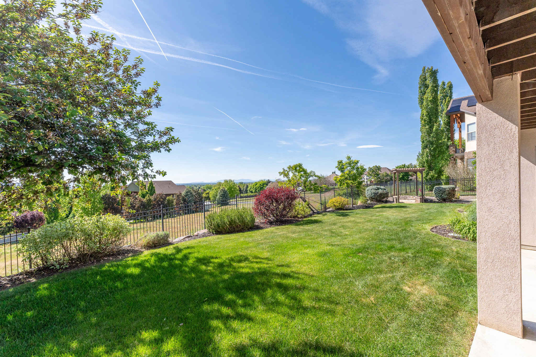 Expansive green lawn surrounded by varied plantings and a black metal fence, with a wooden pergola structure