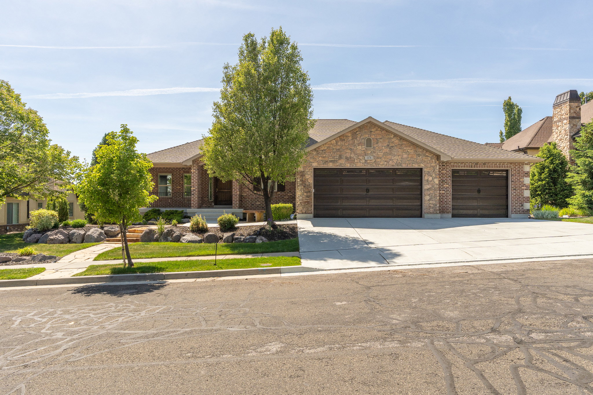 Brick and stone veneer exterior with a multi-car garage, concrete driveway, and a front yard featuring mature trees and decorative rock landscaping
