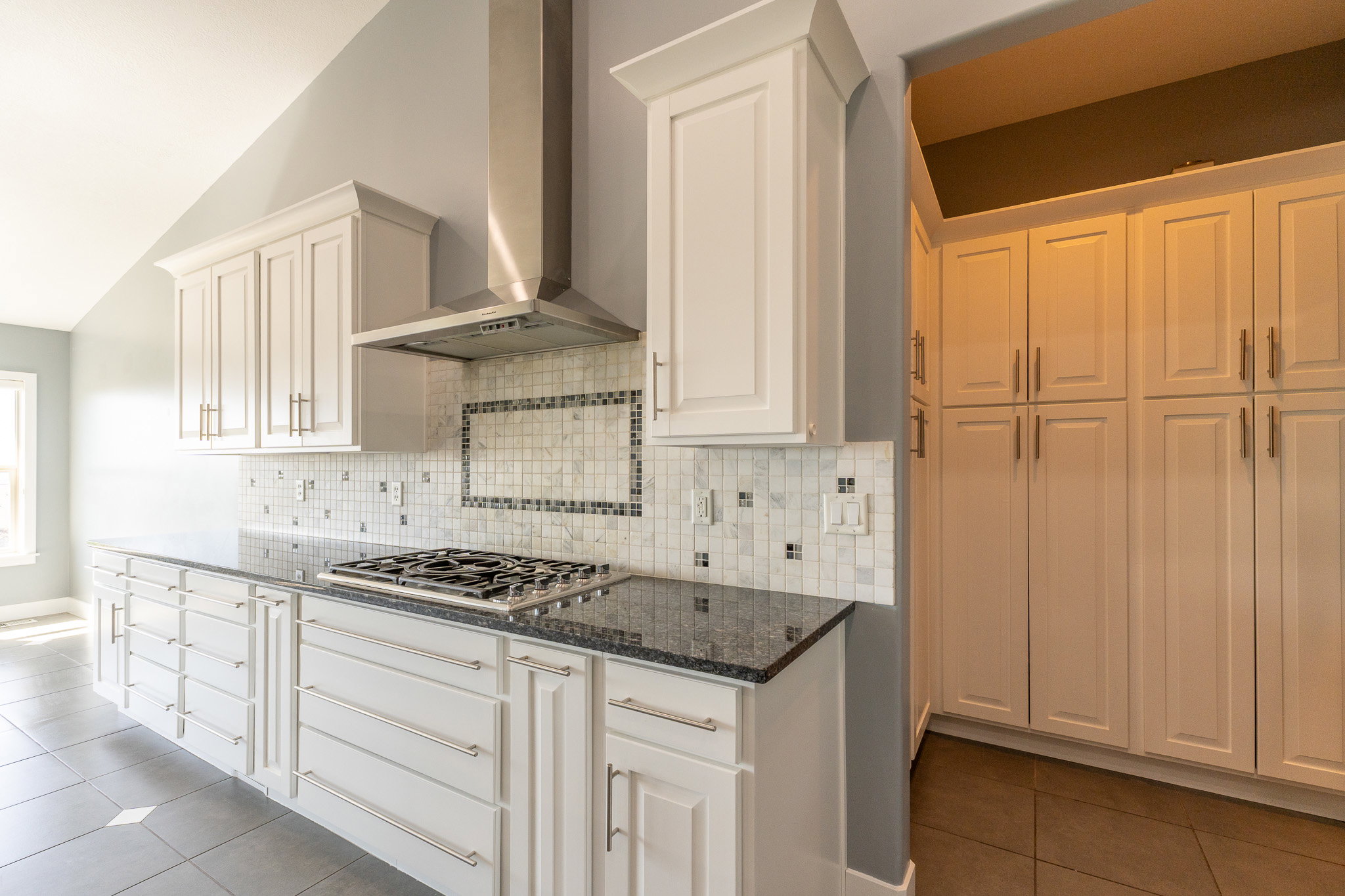 Modern kitchen featuring white cabinetry, dark stone countertops, a gas cooktop with a stainless steel range hood, mosaic tile backsplash, and extensive pantry storage